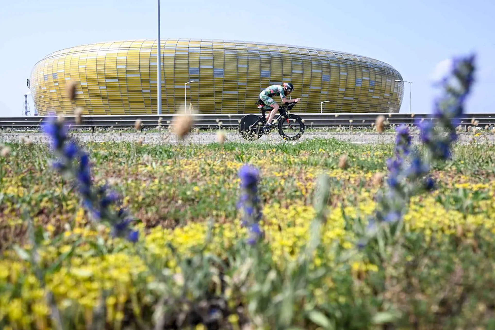 The image shows a person riding a bicycle on a road. In the background, there is a large, distinctive yellow stadium with a unique design. The foreground features some flowering plants, adding color and depth to the scene.