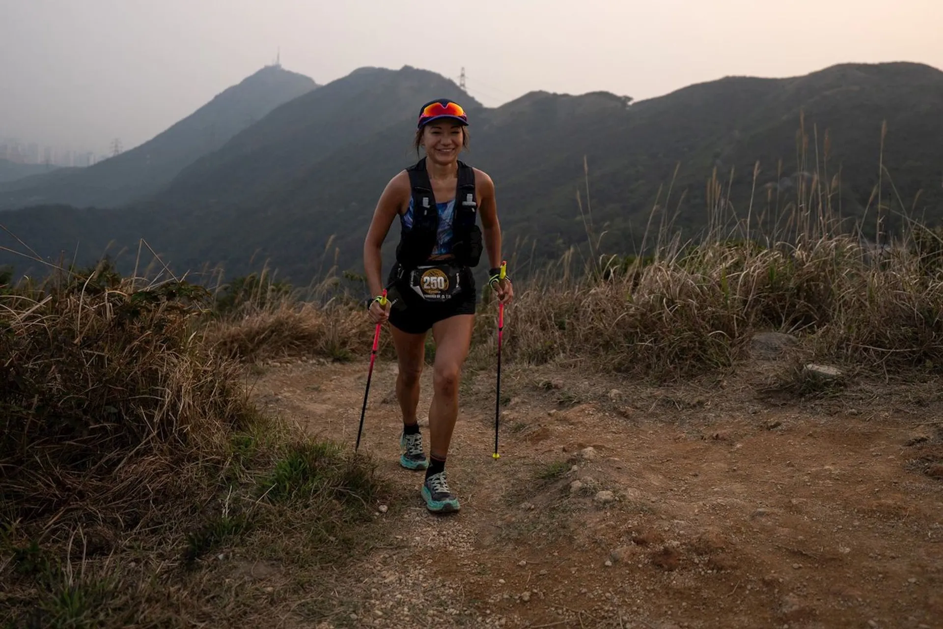 The image shows a person trail running on a dirt path in a mountainous area. The runner is using trekking poles and wearing athletic gear, including a race bib. The background features hills and a hazy sky.