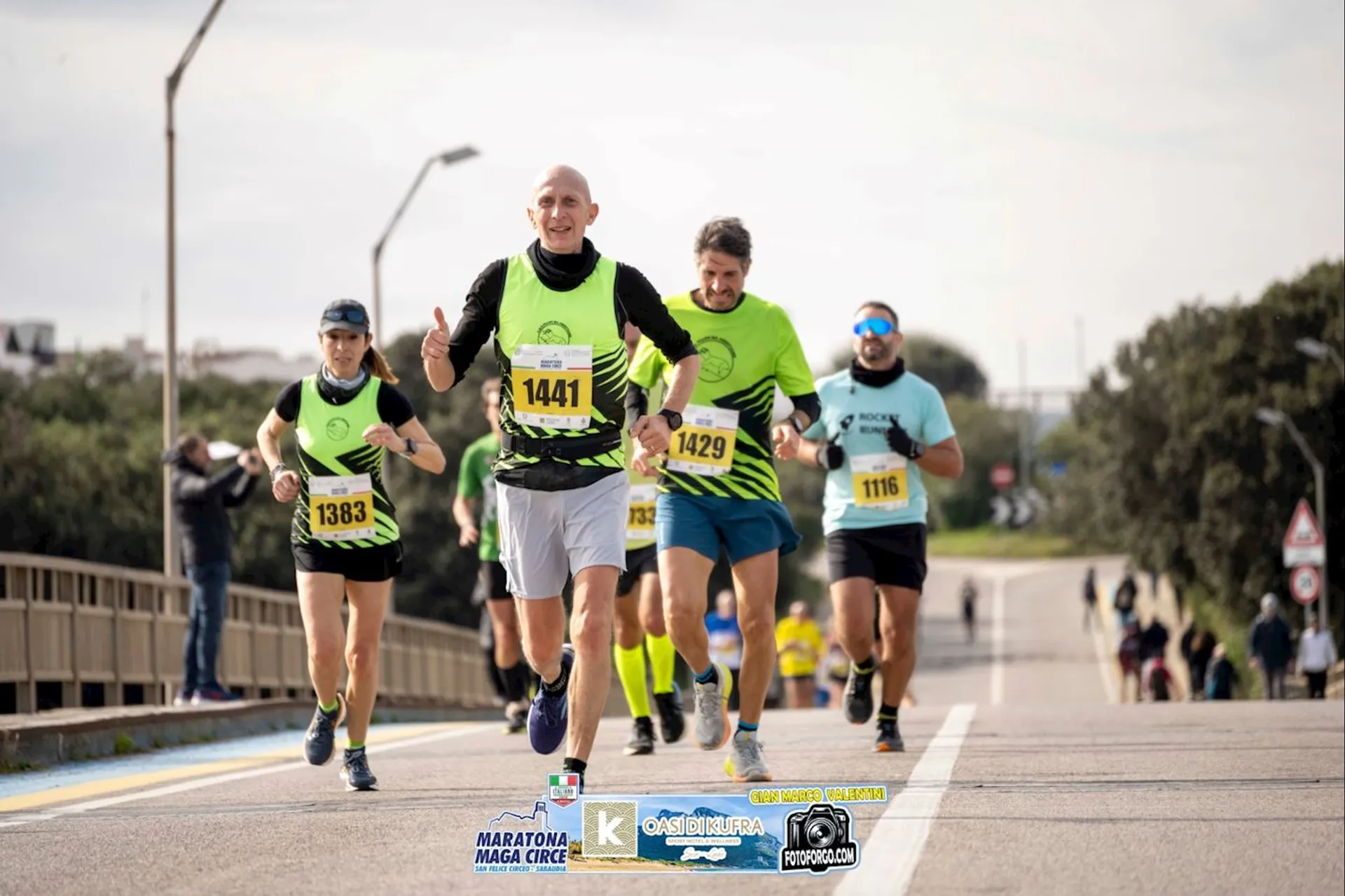 The image shows a group of people running in what appears to be a race or marathon. They are wearing athletic gear and numbered bibs. The background features a road and some signs indicating an organized event. The presence of logos suggests it's part of a marathon event.