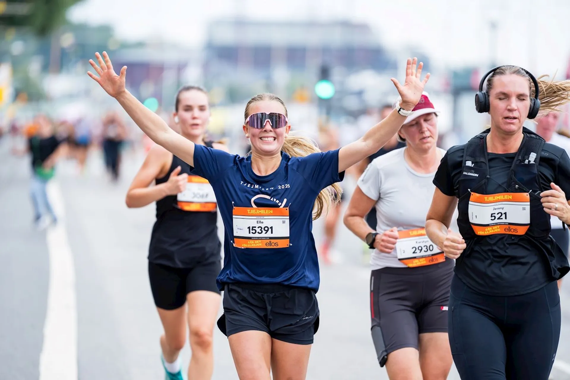The image shows a group of people running in a road race. The participants are wearing athletic gear and race bibs with numbers. One woman in the center has her arms raised and appears to be celebrating. Other runners are visible in the background, some wearing headphones. It seems to be taken during an organized running event or marathon.