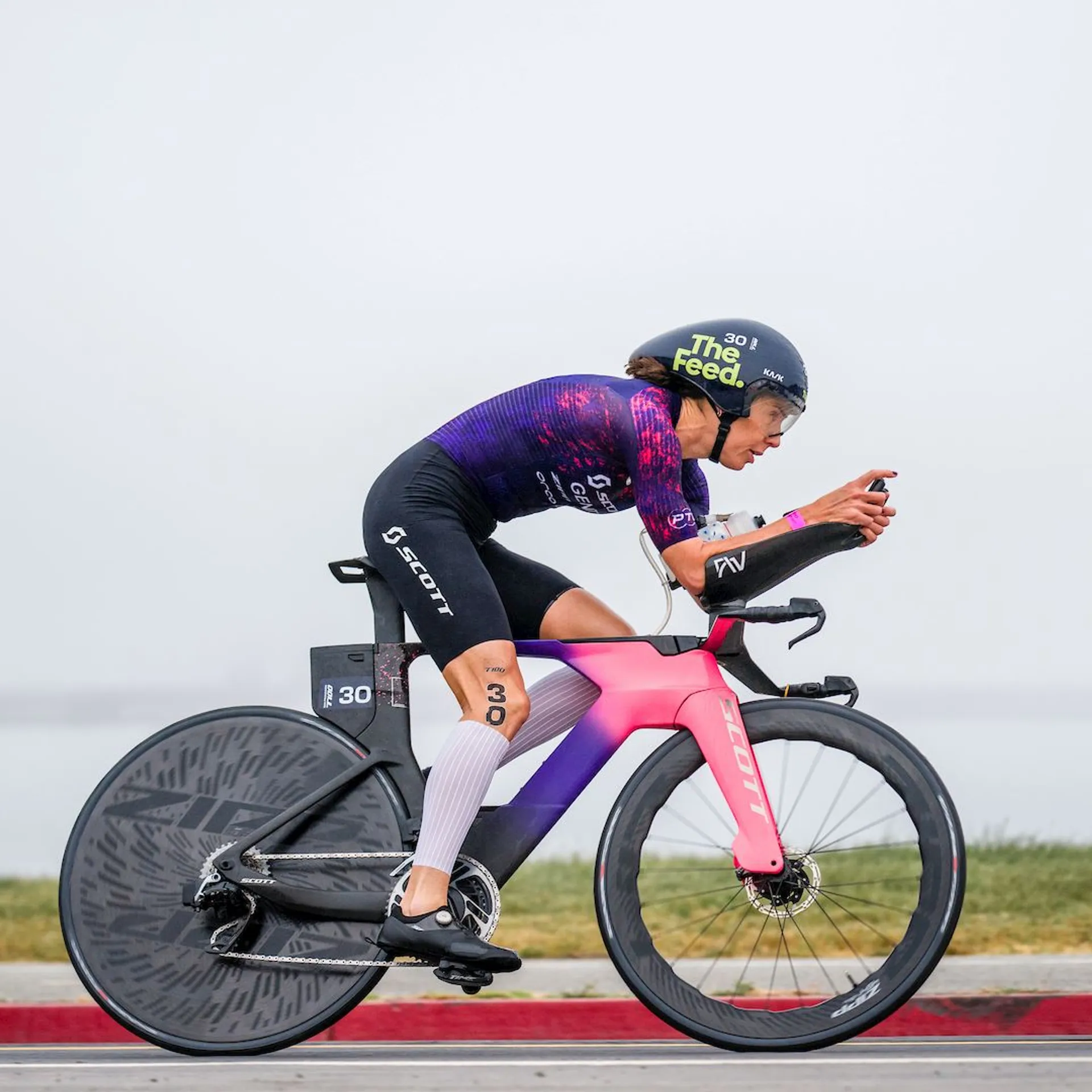 In the image, you see a cyclist in a race position riding a time trial bike. The cyclist is dressed in a professional, tight-fitting cycling suit with sponsor logos and is wearing a time trial helmet, which is aerodynamically shaped to reduce wind resistance. The bike is equipped with aero bars, which the cyclist is using to maintain an aerodynamic position, and it has disc wheels, also designed to minimize drag. The scene seems to be during a competitive cycling event, possibly a triathlon or time trial race, judging by the number "30" and race details on the athlete's arm and helmet. The environment appears to be overcast, and this is an outdoor setting, possibly near a body of water, given the hint of