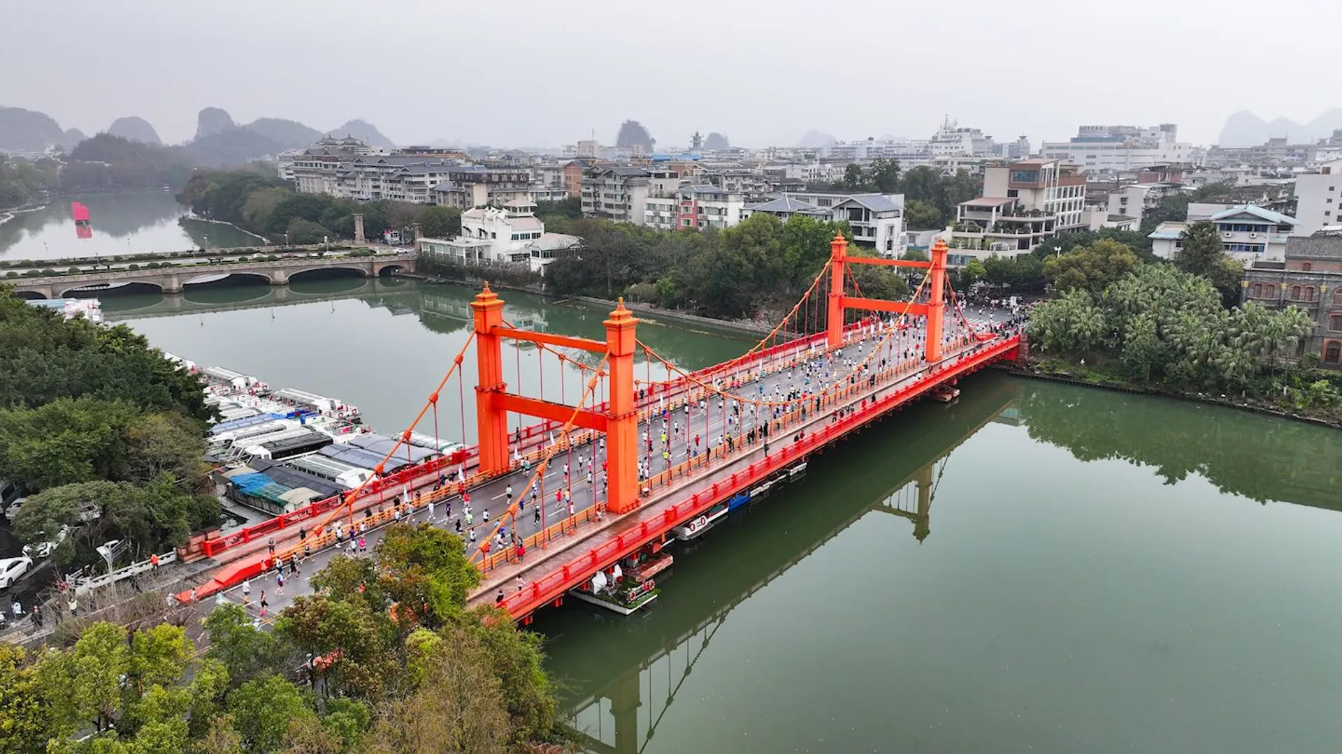 The image shows a vibrant orange suspension bridge spanning over a river, surrounded by a cityscape. The bridge appears busy with people walking across it. There are buildings on either side of the river, and in the background, there are tree-covered hills. The scene suggests an urban area with a mix of natural and built environments.