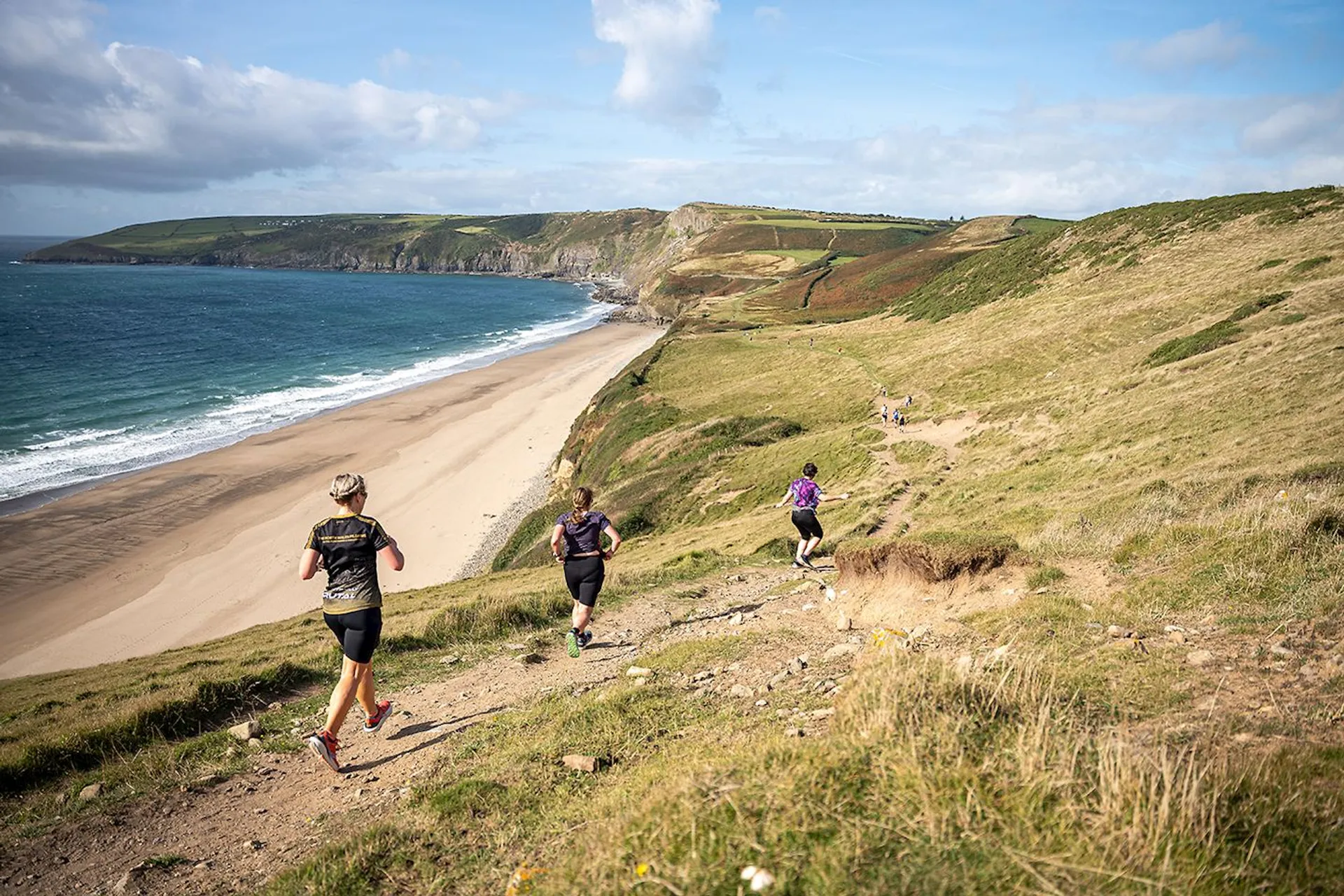 The image shows several people running on a trail along a scenic coastal area. The scene includes grassy hills, a sandy beach, a body of water, and a clear sky with some clouds. The runners appear to be participating in an outdoor activity, enjoying the natural landscape.