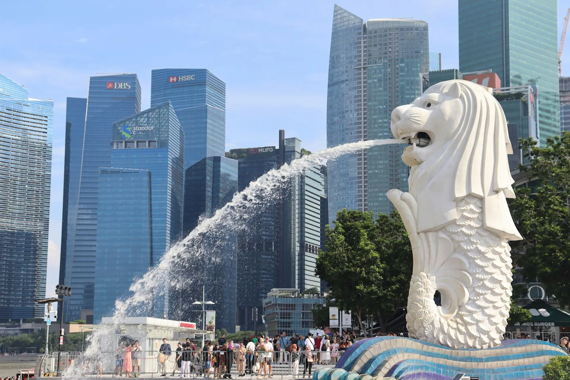 The image shows the Merlion statue in Singapore, a famous landmark that has the body of a fish and the head of a lion. It's located in Merlion Park, with the Singapore skyline visible in the background, featuring several high-rise buildings. The statue is spouting water from its mouth into the bay.
