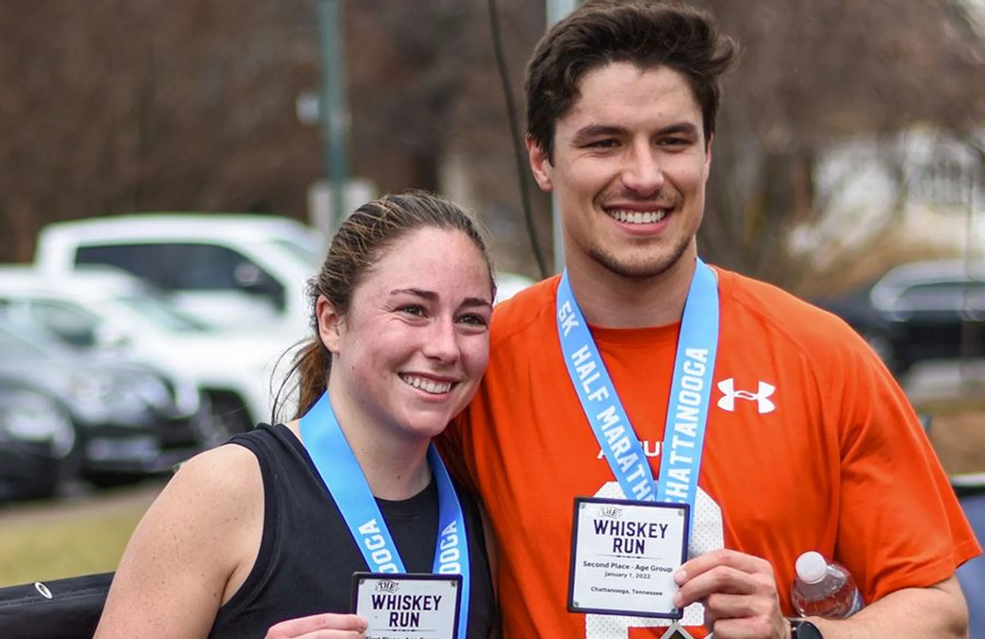In the image, you can see two individuals smiling and posing for the photo. They appear to be at a running event, as they are both wearing medals around their necks that suggest they have completed a race, possibly named "Whiskey Run," as indicated by the text on their medals and the race bib one of them is holding. The bib also includes "Chattanooga," which might suggest the location of the event. They seem to be dressed in athletic gear suitable for running, and the surroundings include leafless trees and parked cars, which indicates the photo may have been taken in a cooler season or a location with a temperate climate. Both seem to be in good spirits and are likely proud of their achievement at this event.