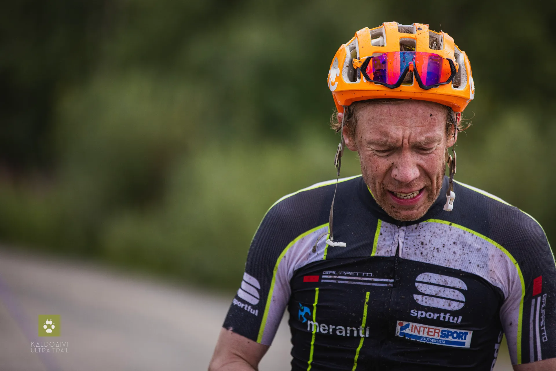 The image shows a male cyclist who appears to be in the middle of a strenuous ride, as suggested by the mud on his face and clothes, and his expression of exertion or fatigue. He is wearing a helmet and sports attire, which includes a black jersey with various sponsor logos, indicating that he might be participating in a competitive cycling event or is at least engaged in serious cycling activity. The background is blurred but suggests a natural outdoor setting, consistent with a trail or road commonly used for cycling.