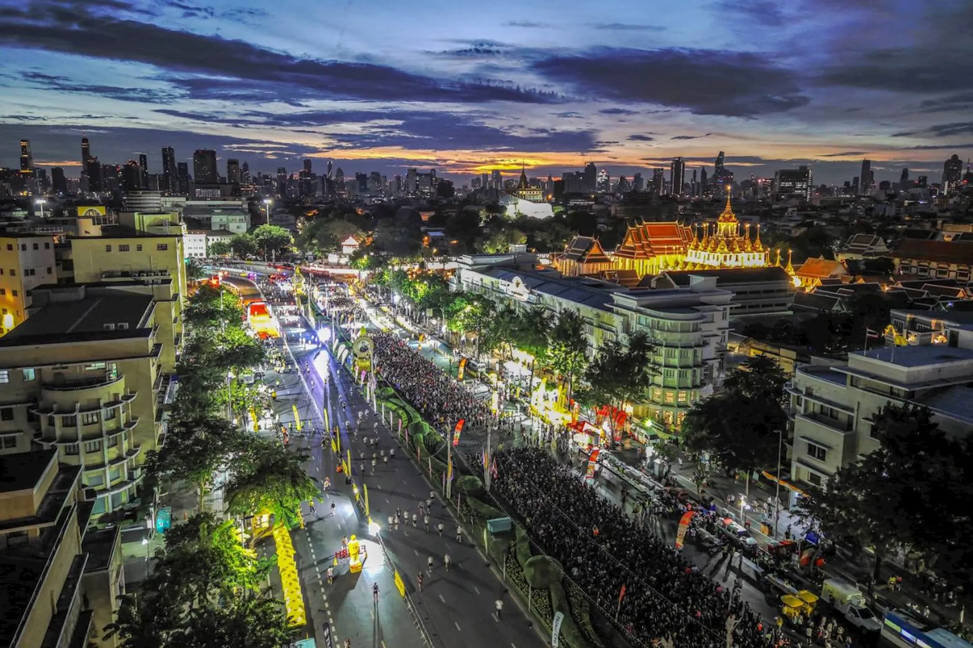 This image shows an aerial view of a vibrant, bustling cityscape at sunset or dusk. The scene is lively with bright lights and crowds along the streets, suggesting a festival or large event. There's a mix of modern and traditional architecture, with a notable illuminated building with spires in the midground. The skyline in the background features tall buildings under a striking, colorful sky.