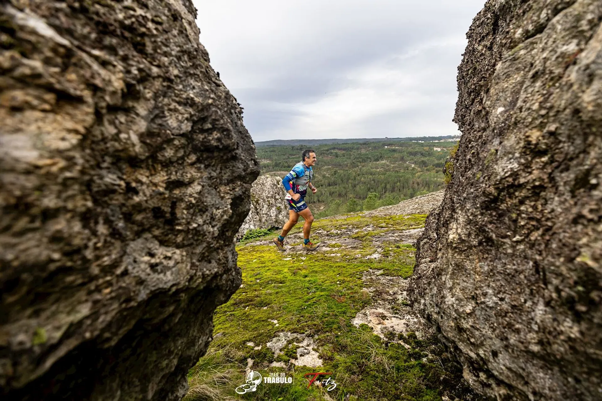 The image shows a person running through a rocky terrain, possibly participating in a trail running event. The runner is surrounded by large rock formations and greenery, with a landscape view in the background. The person is wearing athletic gear and carrying a backpack.