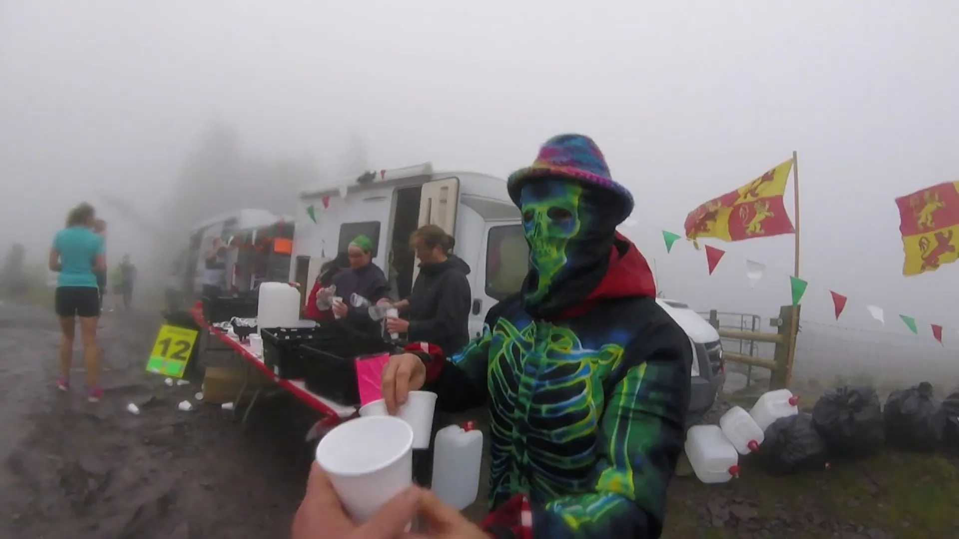 The image shows a person wearing a colorful skeleton costume and mask, handing out a cup. They are at an outdoor event with a festive setup, including flag bunting and tables with supplies. Other people are in the background, some of whom are possibly participants in an athletic event, given the nature of the setup. The surroundings are foggy, and there are several jugs and plastic bags visible.