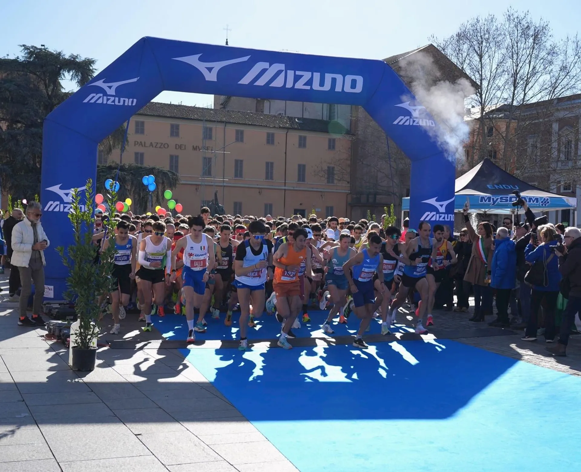 The image shows a group of runners starting a race under a blue arch with the Mizuno logo. There are spectators gathered around the starting line, and a building and some trees are visible in the background. The atmosphere appears to be energetic and lively.