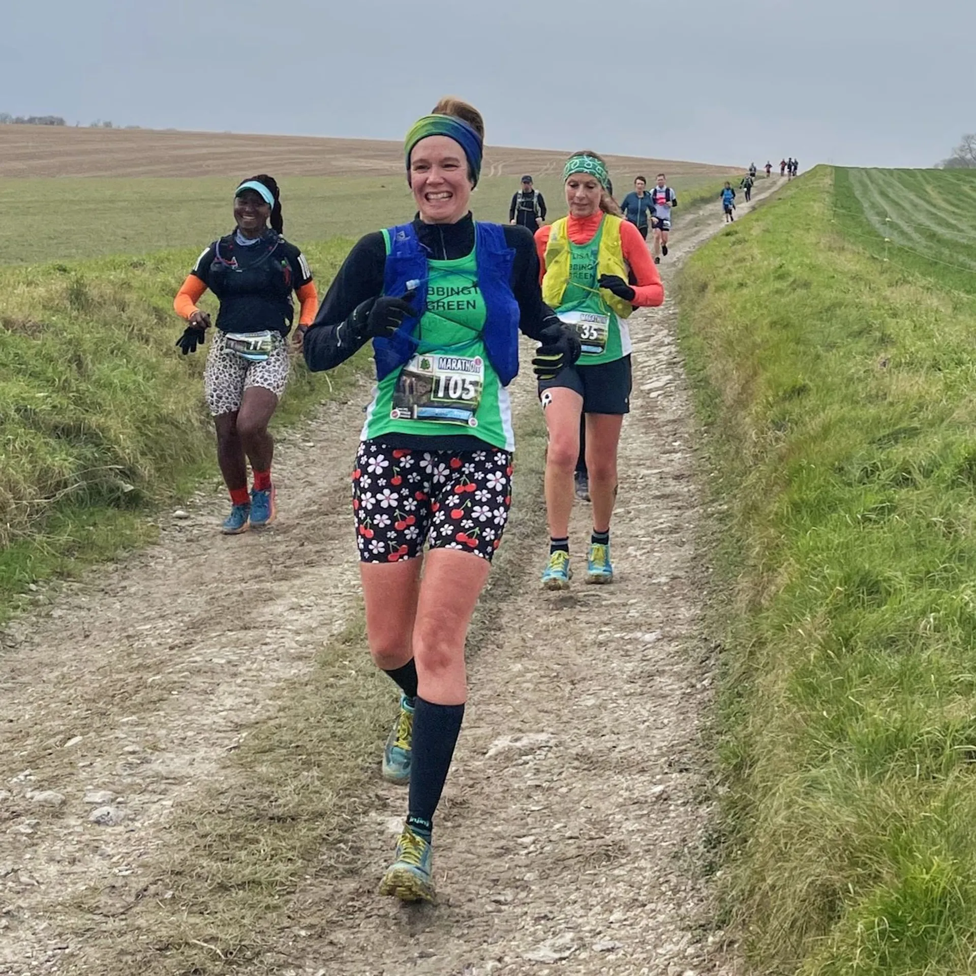 The image shows a group of people running on a dirt path in an outdoor setting. They appear to be participating in a race or marathon. The environment looks rural, with grassy areas and a slightly overcast sky. The runners are wearing bib numbers and various athletic clothing.