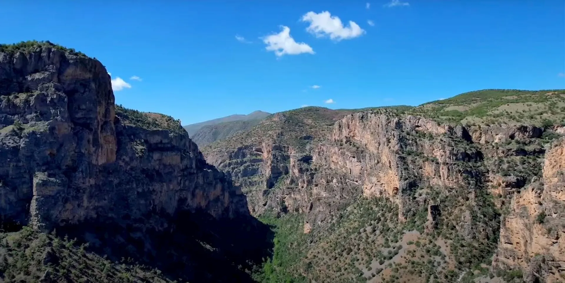 The image shows a dramatic landscape featuring rugged cliffs and a deep canyon. The terrain is mountainous with steep rock faces and some vegetation on the slopes. The sky is mostly clear with a few sparse clouds, highlighting the natural beauty of the canyon area. It seems to be a sunny day and the viewpoint suggests the photo may have been taken from a higher elevation, possibly from a vantage point on the opposite side of the canyon or from an aerial perspective like a drone.