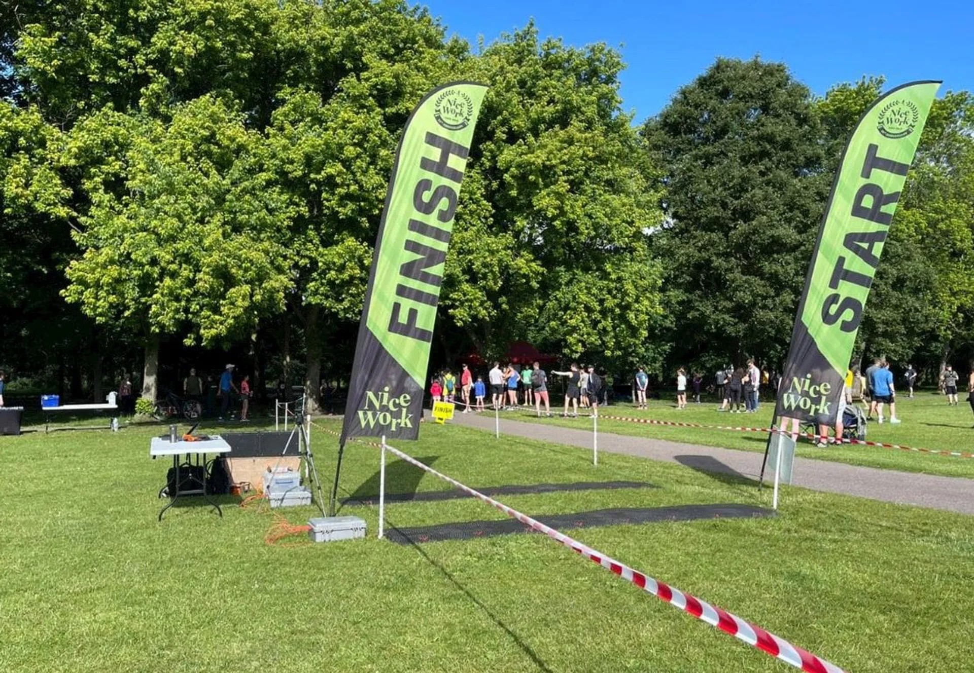 The image depicts an outdoor event, likely a race or fun run, given the presence of "Start" and "Finish" banners. The picture is taken on a sunny day with clear blue skies. There are a number of people gathered around, some of whom are participants standing behind the "Start" line preparing to begin the event.

To the left, there is a table set up with various items on it, which might be used for registration, timing, or providing water and snacks to participants. The area is demarcated by red-and-white striped tape, and there is a clear path designated for runners to follow. The green banners have the text "Nice Work" on them, suggesting encouragement or perhaps the name of the event organizer.

The