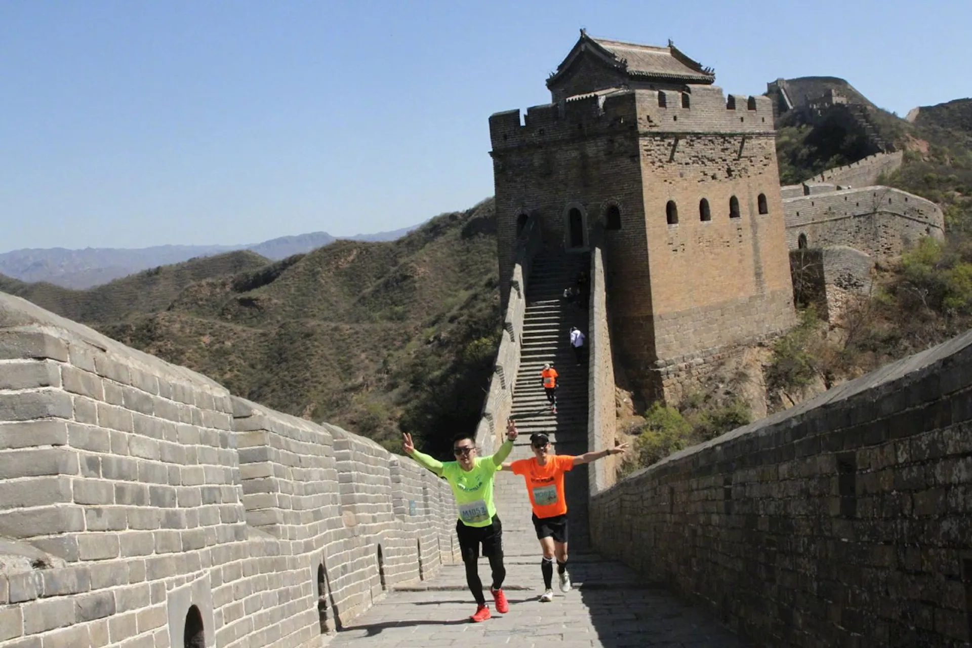 In the image, you see two individuals running along the Great Wall of China. They appear to be in active wear suitable for jogging or exercise, suggesting that they might be participating in a run or some form of a marathon that takes part on the Great Wall. The wall itself is winding along the mountainous landscape with watchtowers and is a historical fortification and one of the most famous landmarks in China. The weather is clear and it looks like a sunny day, ideal for outdoor activities.