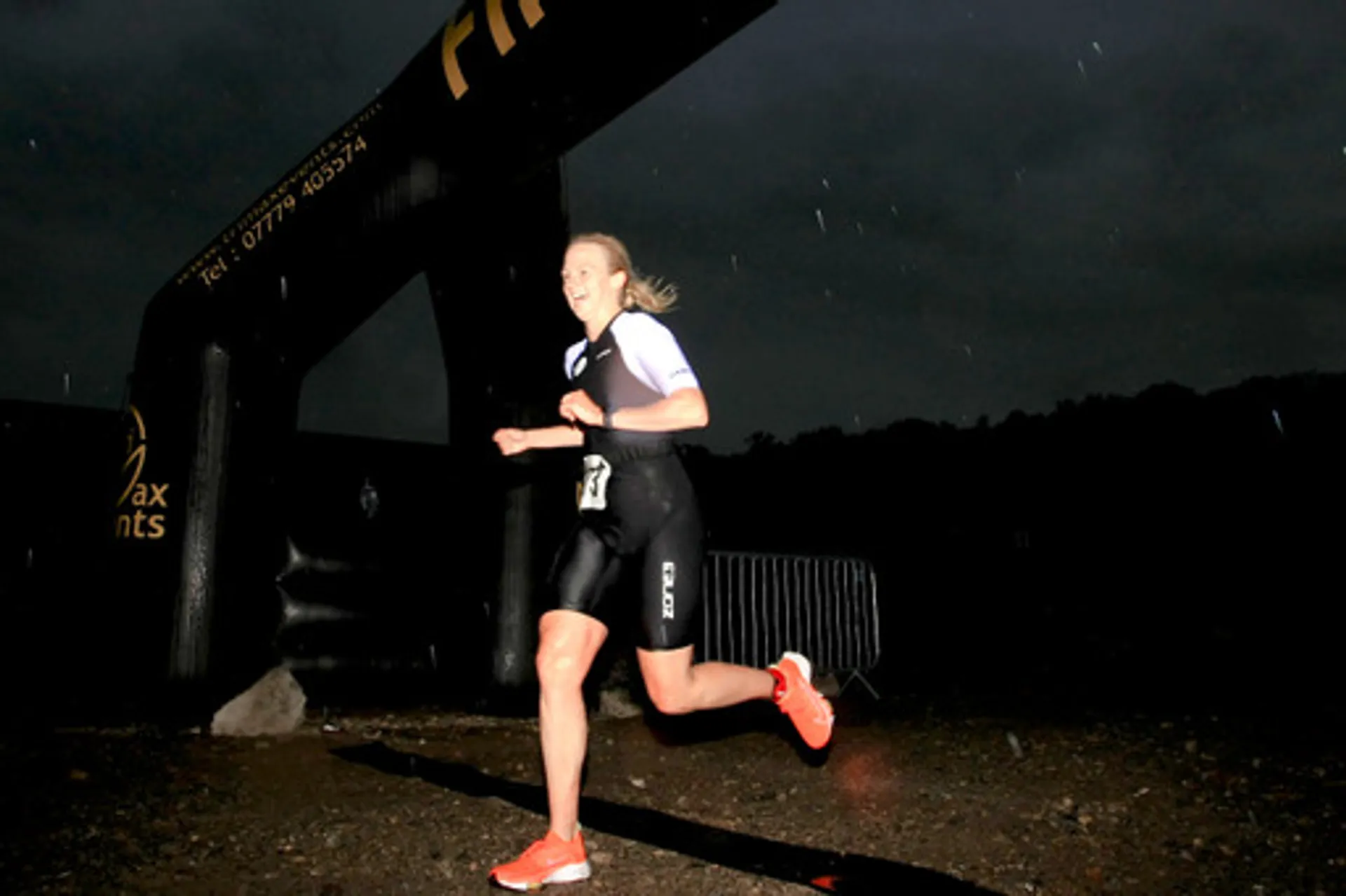 The image depicts a woman who is running at night or during low light conditions. She appears to be in an athletic outfit suitable for running, with a visible race number on her thigh, suggesting she might be participating in a race or fitness event. The woman is smiling and looks energetic as she runs past a structure that seems to be a part of the race setup, possibly the start or finish line, which is marked by an inflatable arch with some text on it. The low light and visible raindrops or moisture suggest the event is taking place outdoors in the evening or night. The scene captures a sense of motion and the setting is somewhat moody due to the darkness and weather conditions.