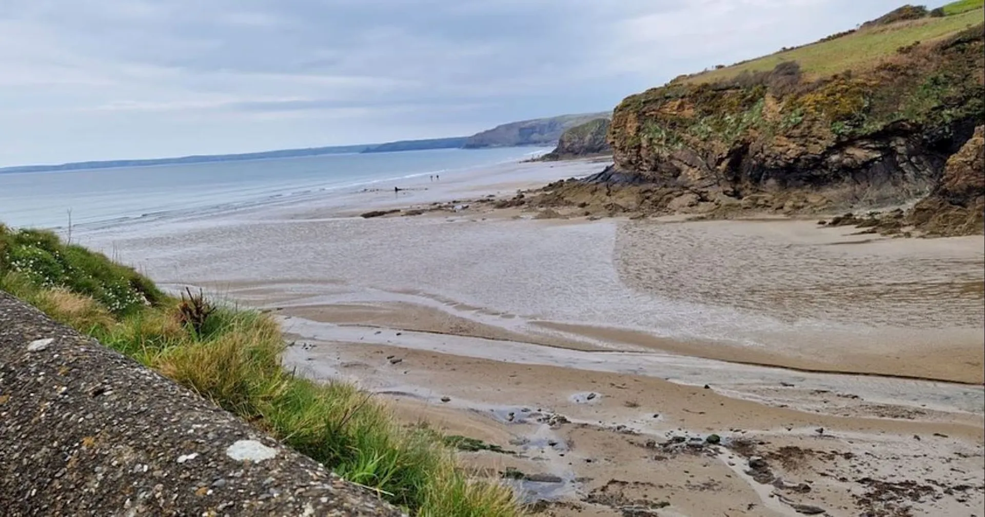 The image displays a tranquil coastal scene, likely at low tide given the wide expanse of exposed wet sand. The coastline extends into the distance, framed by cliffs or hills on the right. Vegetation can be seen atop these elevation changes, suggesting a natural and possibly rural area. The ocean is calm, and the sky is partly cloudy, indicative of fair weather. There are a few people in the distance enjoying the beach, appearing as small figures in the vast landscape. A barrier or wall is visible in the foreground on the left, possibly a walkway or promenade alongside the beach.