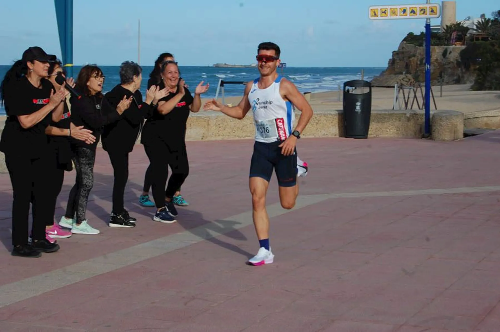 The image shows a runner participating in a race along a coastal path. The runner is smiling and wearing athletic gear, including a bib number. A group of people on the side is clapping and cheering for the runner. The setting is near the ocean, with a beach and some trees in the background.