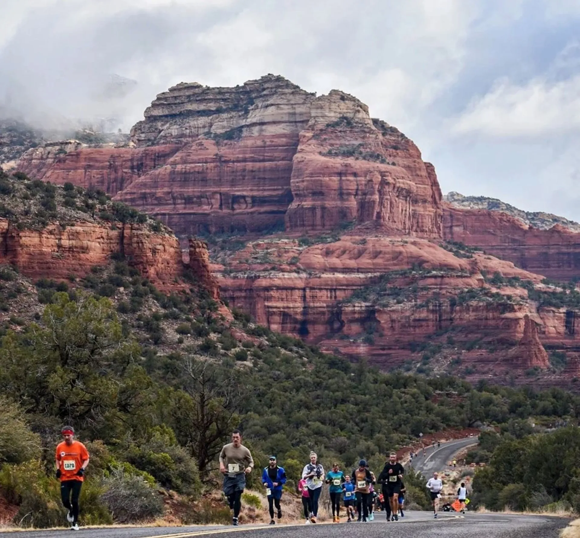 This image shows a group of people running a race on a road through a picturesque desert landscape. In the background, there are impressive red rock formations and lush greenery, suggesting it's in a location with natural features like those found in places such as Sedona, Arizona. The sky is cloudy, adding to the dramatic scenery.