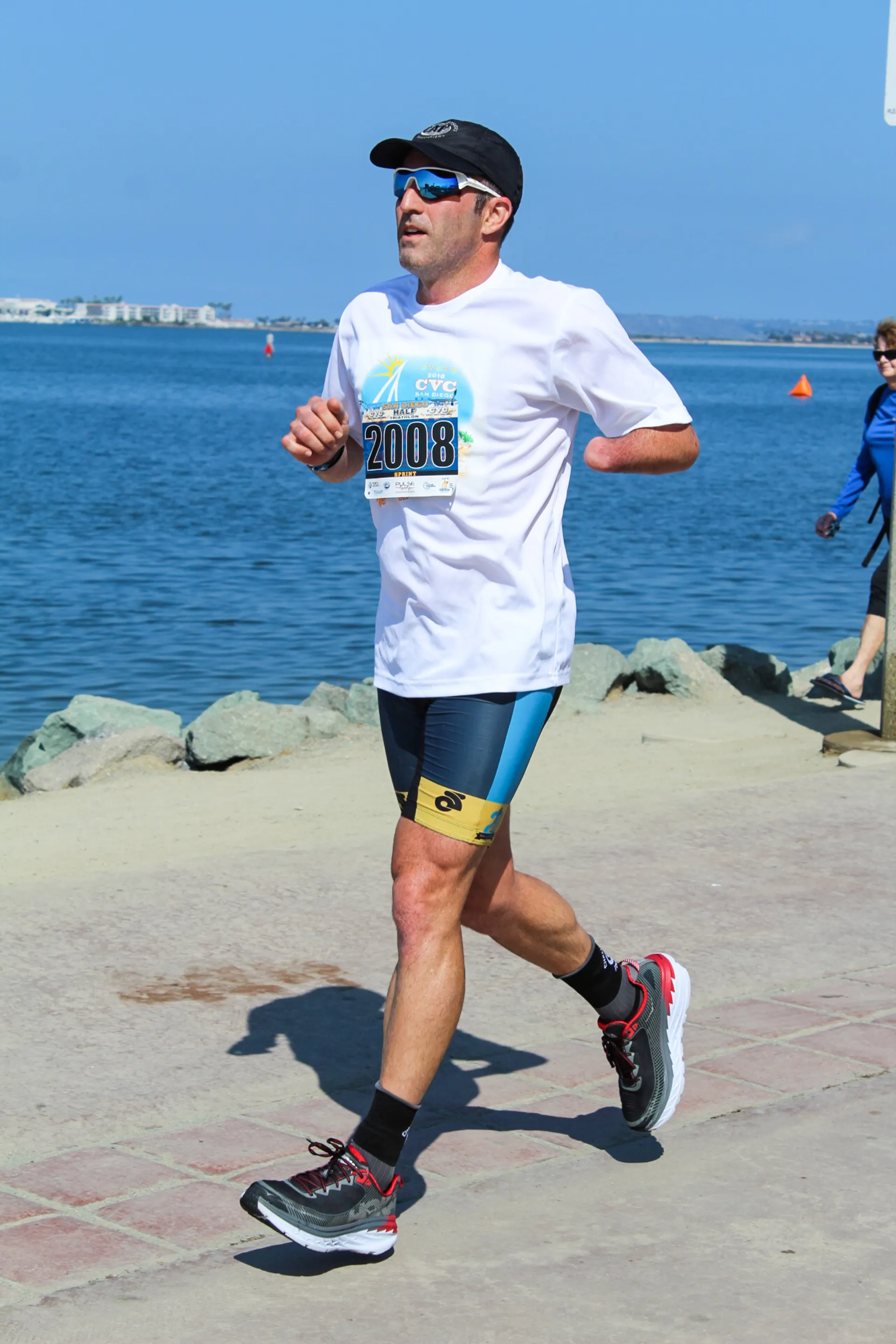 The image shows a man participating in a running event. He is wearing a white t-shirt with a race number "2008" on it, dark shorts with a design on the sides, a black cap, and running shoes. He appears focused and is jogging by a body of water with a clear sky above him. There is another participant in the background, suggesting this might be part of a race or marathon. The setting looks like a pleasant, coastal area.