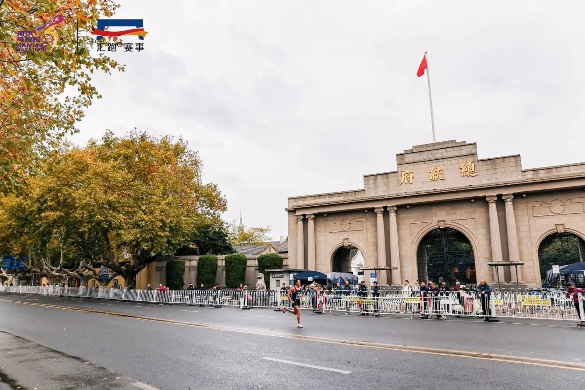 The image shows a street scene with a large building featuring an archway. There's a runner and some people gathered along the street. The building displays a red flag on top and some Chinese characters. Trees are visible, and the sky appears overcast.