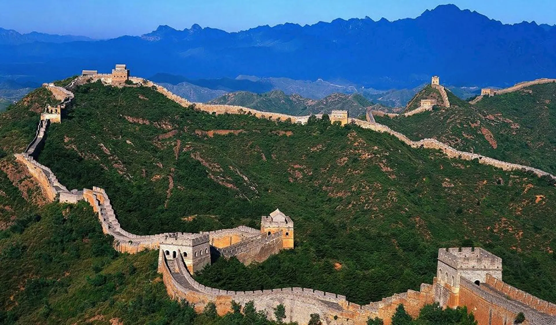 The image shows the Great Wall of China winding through a hilly landscape. The wall is made of stone and brick, and features watchtowers at various intervals. It extends across the ridges with mountains visible in the background.