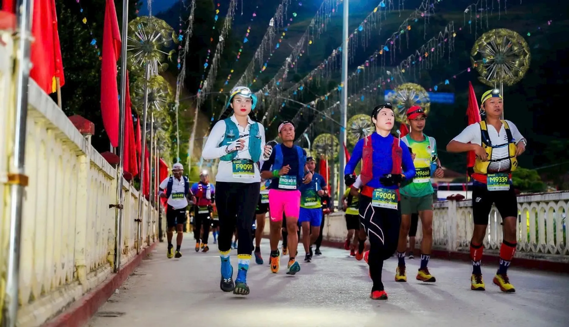 The image shows a group of runners participating in a marathon or similar event. They are running along a decorated path, adorned with lights and festive elements. The runners are wearing athletic gear and numbered bibs.
