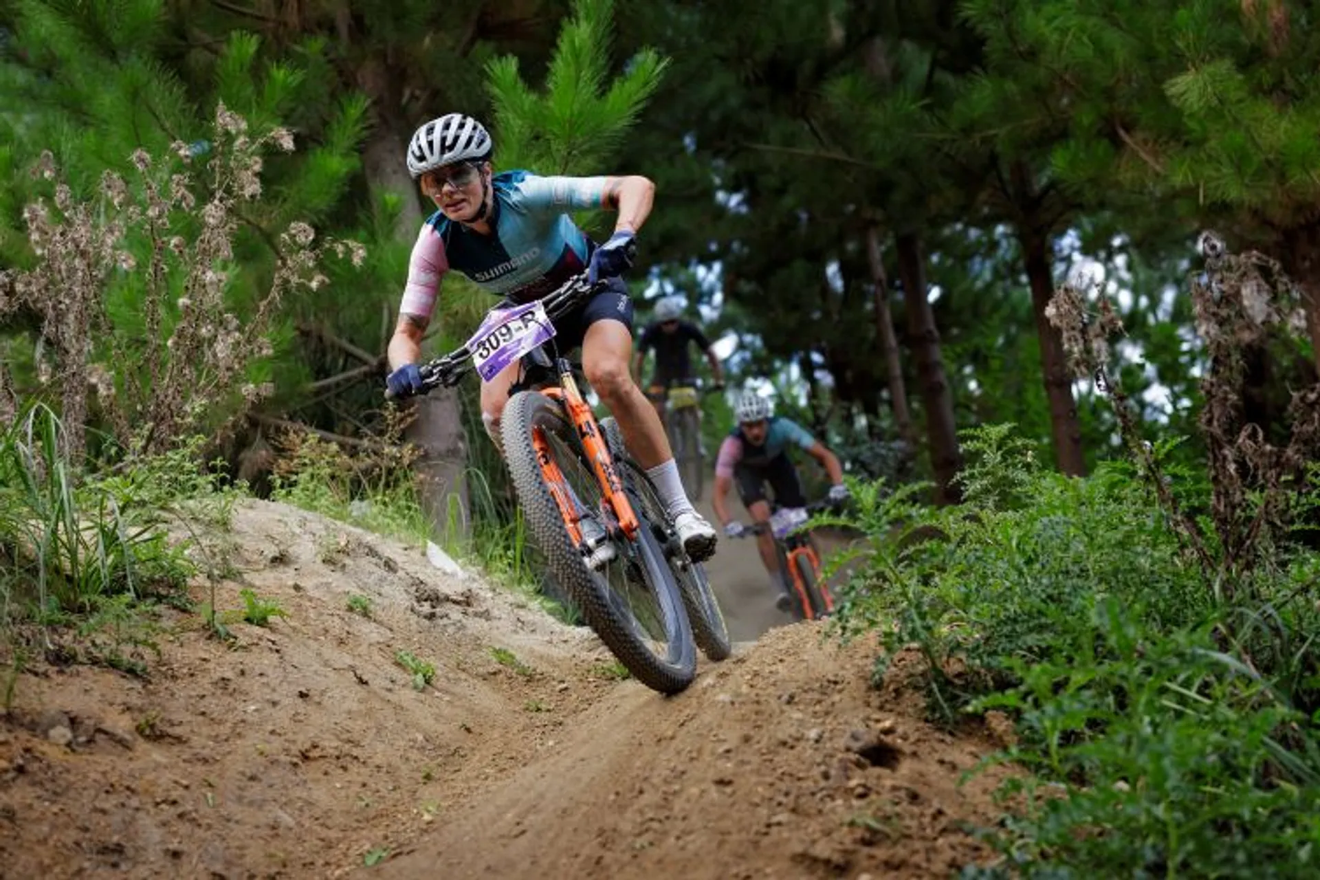 The image shows two mountain bikers riding on a dirt trail in a forested area. The rider in the foreground is wearing a helmet, glasses, and cycling gear. They are navigating a downhill section with some plants and trees around them. The rider in the background is following closely.