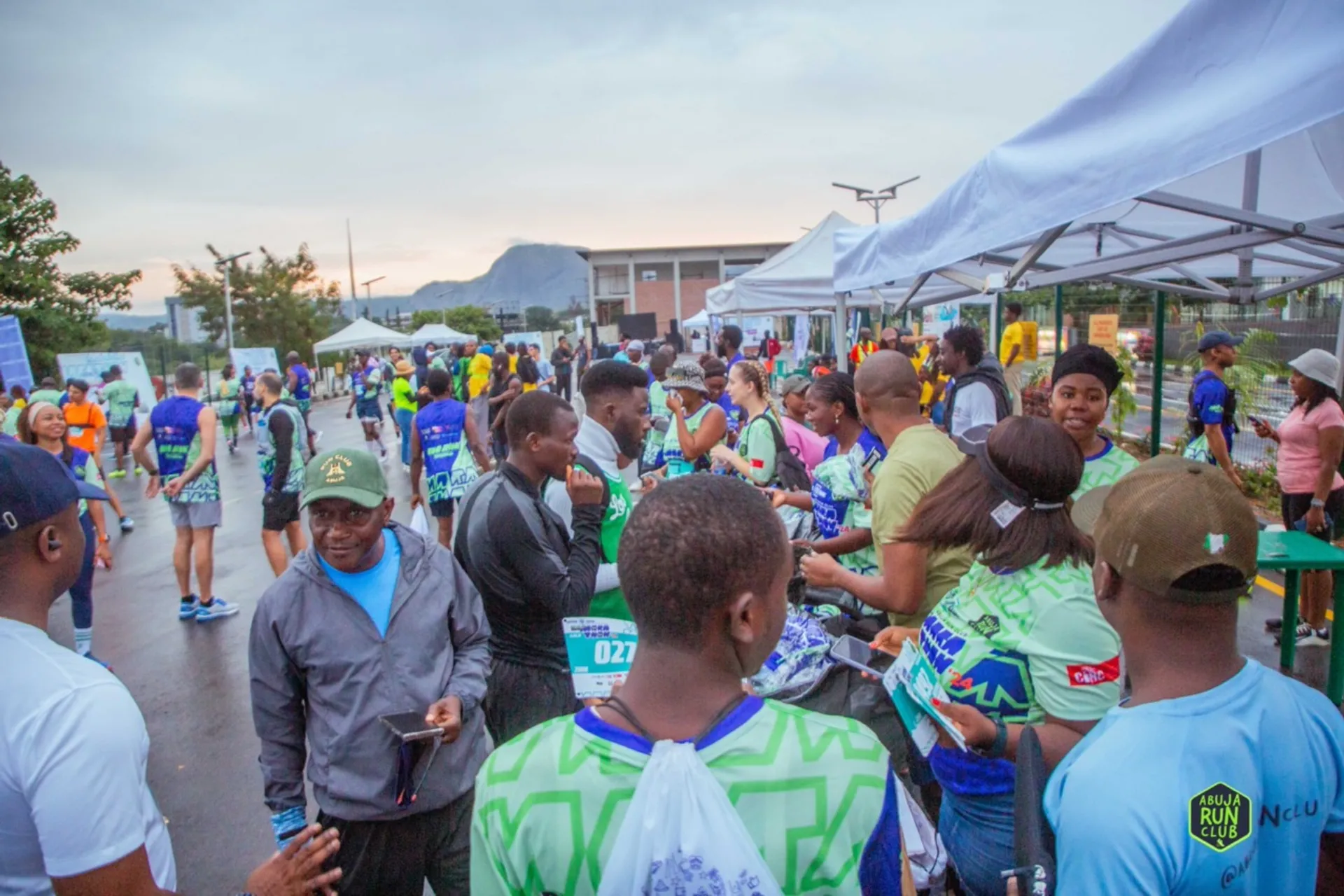 This image shows a gathering of people, likely at an outdoor event, such as a race or marathon. Many of the participants are wearing athletic clothing, some with visible race bibs. There are tents set up in the background, possibly for registration or refreshments. The atmosphere seems active and social, with people engaging with each other.