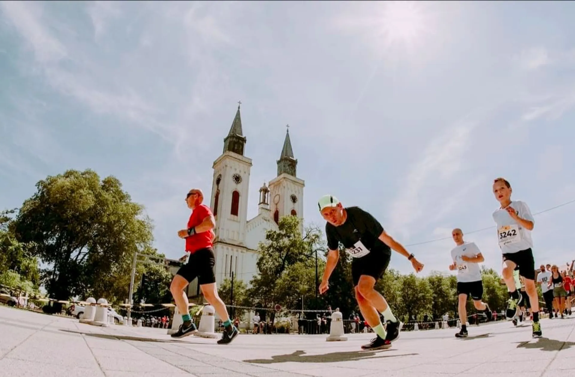 The image shows a group of people running in a race, likely a marathon or similar event. They are in an outdoor setting with a church or cathedral in the background, featuring two tall spires. The sky is clear and sunny, suggesting nice weather.