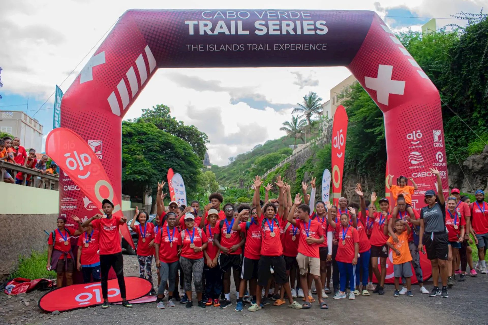 The image shows a group of people gathered under an archway that reads "Cabo Verde Trail Series" and "The Islands Trail Experience." They appear to be participating in an event or race, with many wearing medals and matching red shirts. Some people have their hands raised. There are banners and greenery in the background.