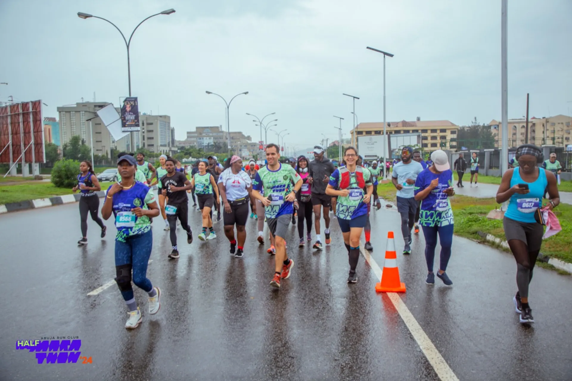The image shows a group of people running in a race on a wet road. They are wearing athletic clothing and numbered bibs. There are traffic cones along the road, and the setting appears to be an urban environment with buildings and streetlights in the background. It's a cloudy day, and the road surface is wet, indicating recent rain.