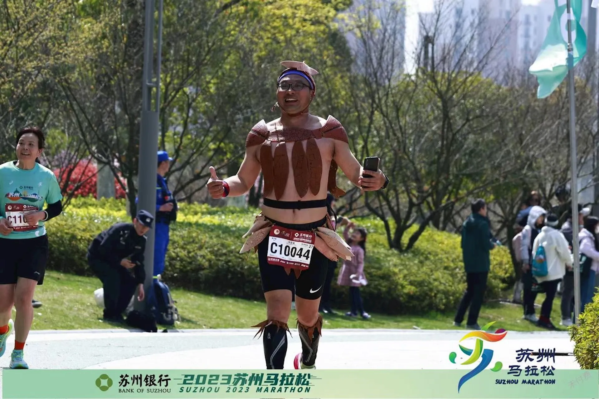 The image shows a person running in a marathon. The runner is wearing a unique costume that appears to be made of items resembling bacon strips. The background has other runners and greenery, and there are logos and text indicating it is the 2023 Suzhou Marathon.