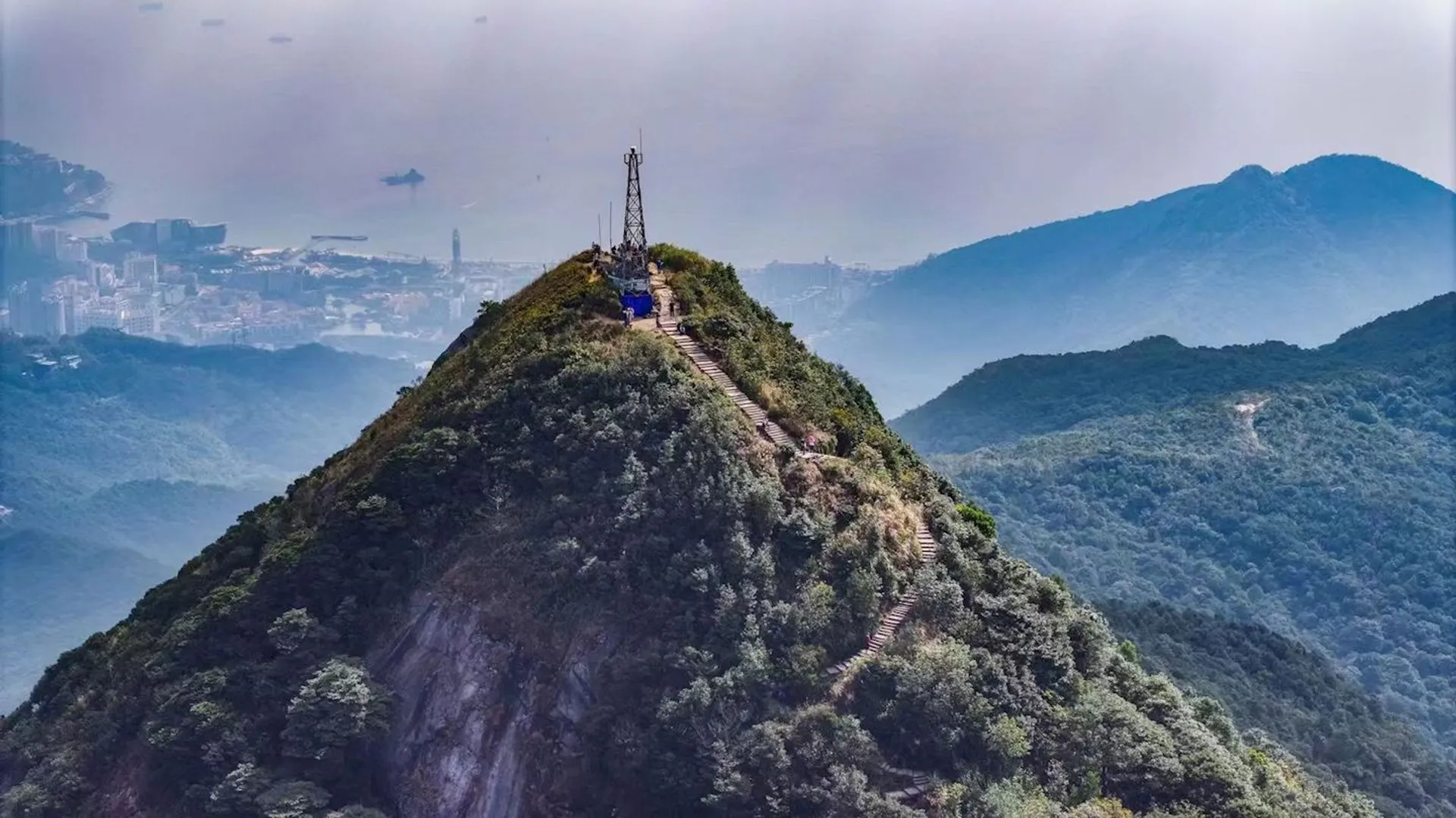 The image depicts a picturesque mountain ridge with a pedestrian pathway or stairs leading up to a summit with a structure that looks like an antenna or observation tower. The surrounding landscape has lush greenery, and there is a layer of mist or fog that adds to the scenic beauty of the view. In the background, you can see more hills or mountains and even part of a cityscape with buildings peeking through the haze, suggesting that this location is near an urban area. The sky appears overcast, and the overall scene conveys a serene and perhaps a slightly mystical atmosphere.