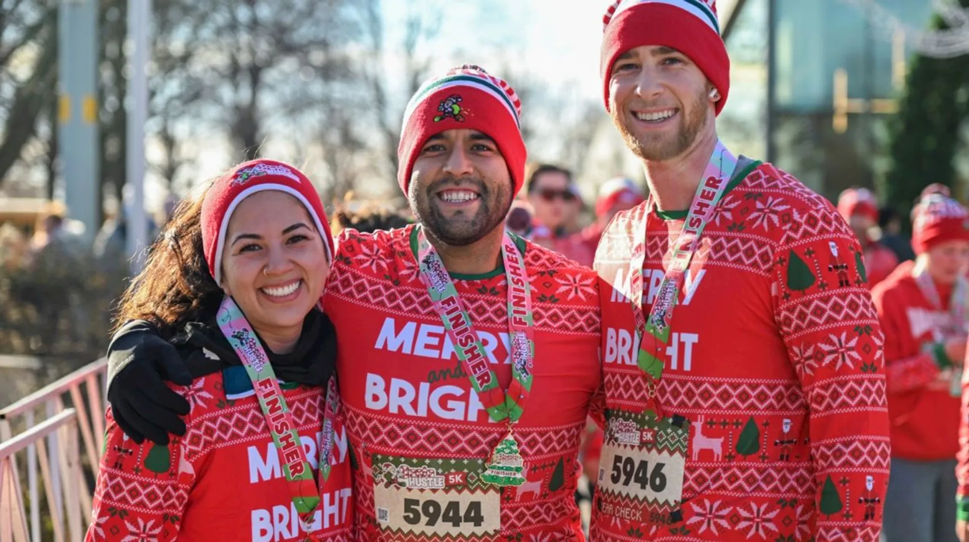 The image shows three people wearing matching festive outfits with red sweaters that say "MERRY BRIGHT" and are decorated with holiday-themed patterns. They are also wearing red and white hats and have finisher medals around their necks, suggesting they participated in a race or event. They appear happy and are posing together outdoors.