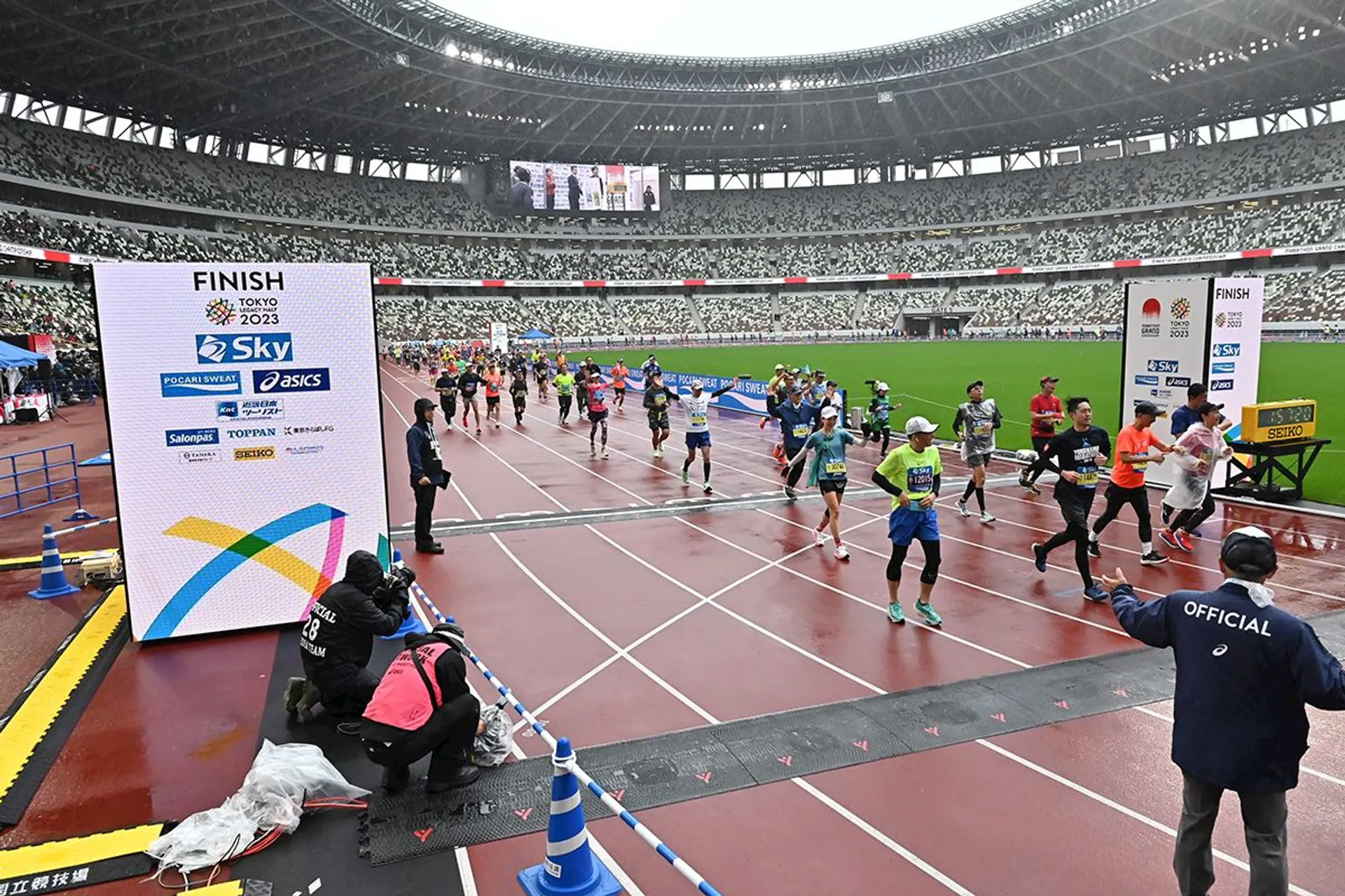 The image shows a group of people running on a track inside a stadium. The runners are approaching a finish line display that reads "FINISH Tokyo 2023" with various sponsor logos beneath it. There are spectators in the stands, and officials are present near the track.