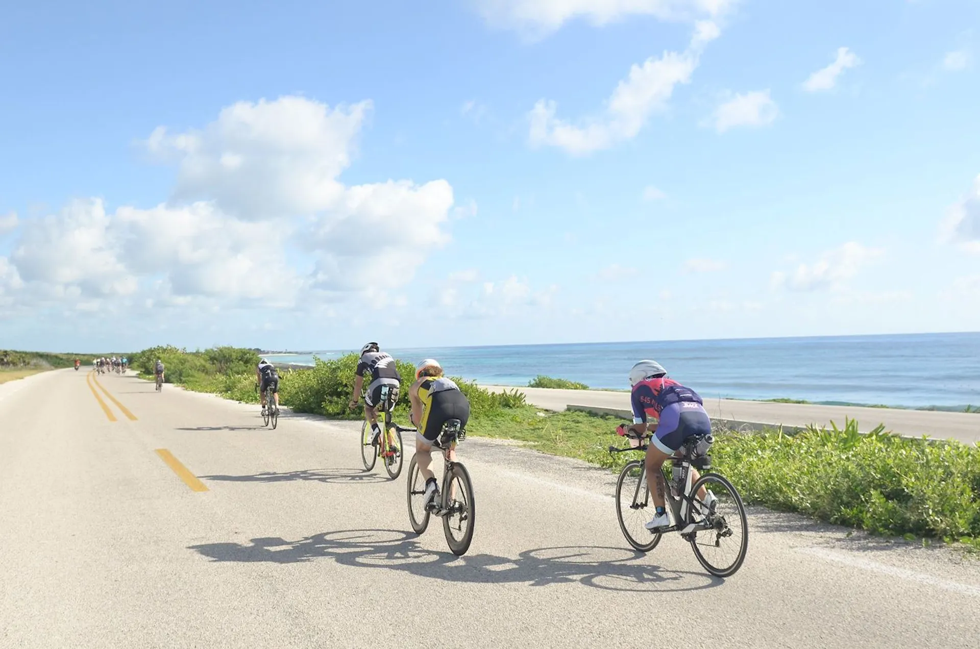 The image shows a group of cyclists riding along a coastal road. The ocean is visible on the right, suggesting that the road may be running parallel to a coastline. The sky is mostly clear with just a few clouds, indicating good weather conditions for cycling. The natural scenery, including the vegetation on the side of the road, suggests a peaceful and scenic route. The cyclists are equipped with road bikes and are wearing athletic gear, helmets, and sunglasses, which are typical for individuals engaged in road cycling or long-distance biking.