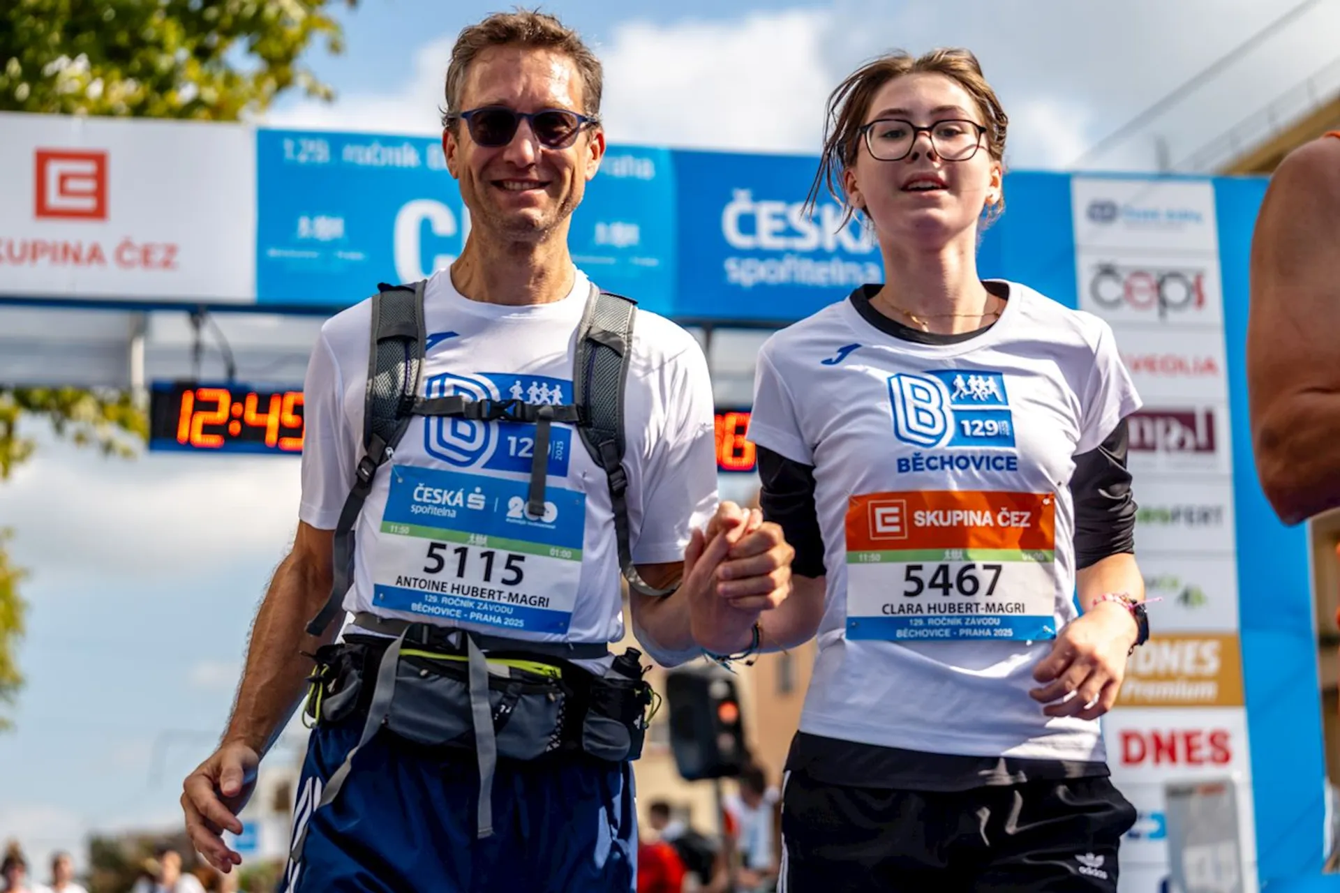 The image shows two people, a man and a woman, participating in a running event. Both are wearing bib numbers and athletic attire. There are banners and signs in the background indicating the event is likely sponsored or organized by "Česká spořitelna" and "Skupina ČEZ."