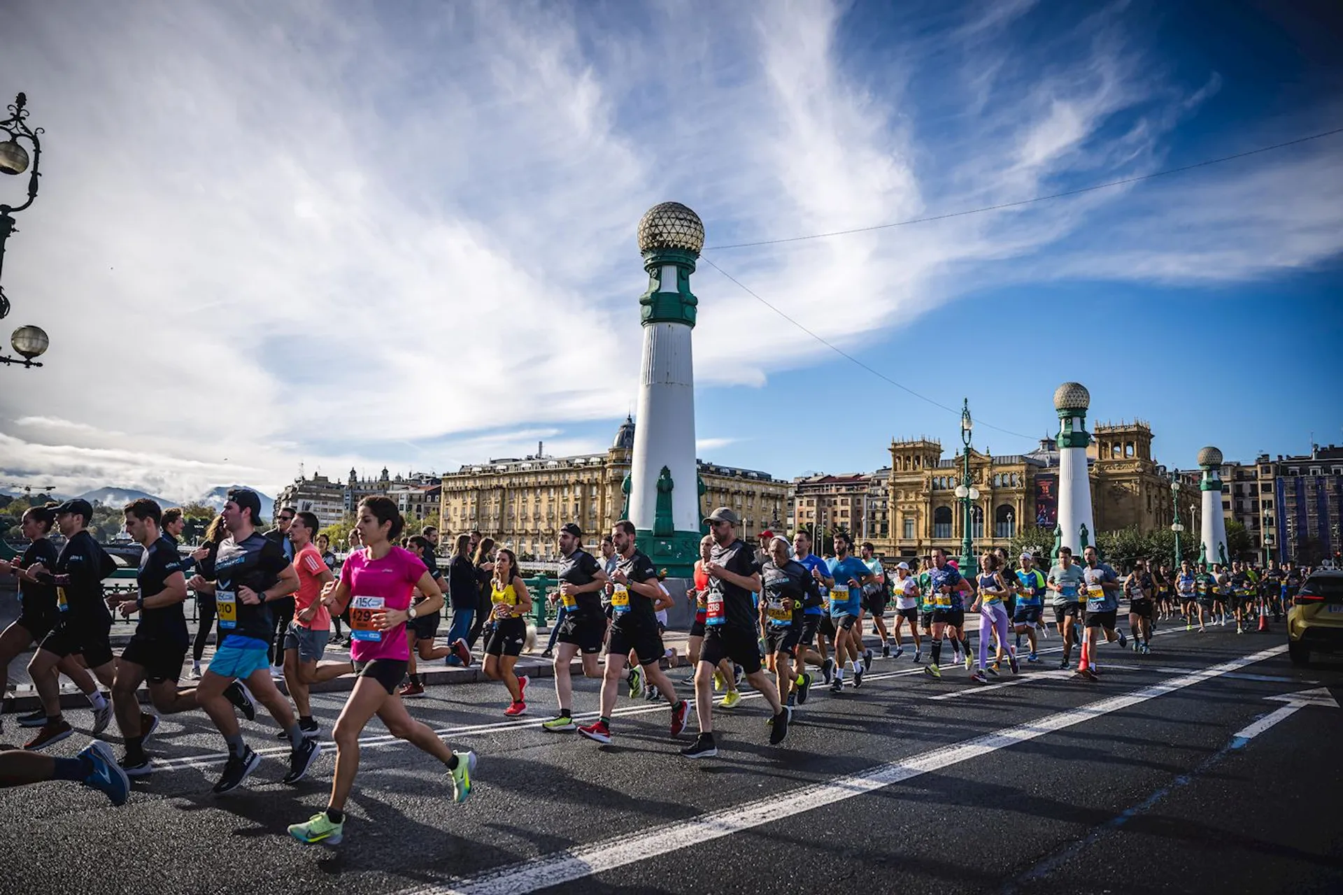 This image shows a group of runners participating in a road race. They are running on a wide street with distinctive architectural features in the background, including large pillars topped with spherical structures. The scene suggests a city marathon or organized running event, and the weather appears to be clear.