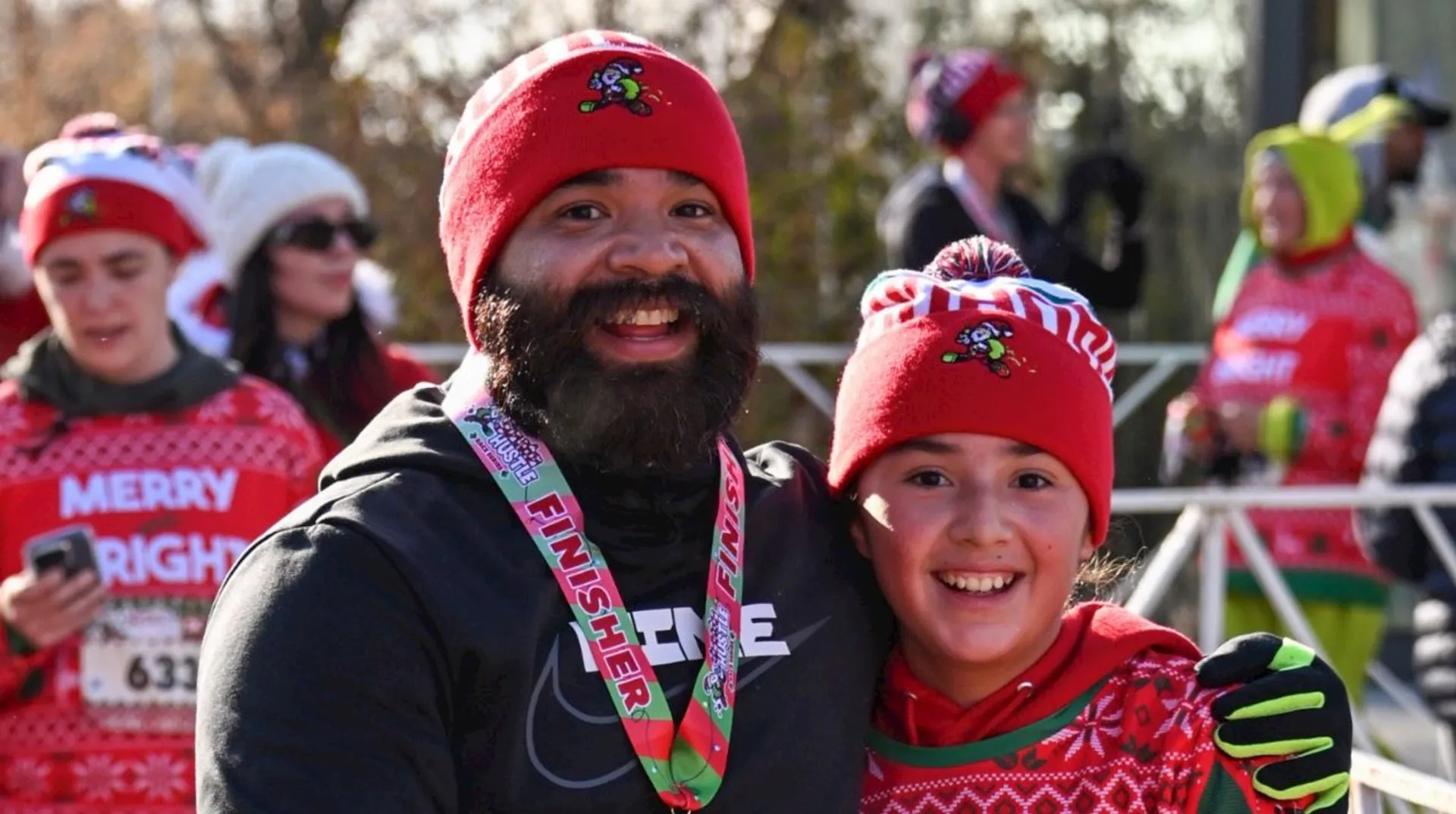 The image shows two people wearing red winter hats and festive clothing, likely participating in a holiday-themed event or race. One is wearing a "Finisher" medal. The background includes other people dressed similarly for the event.