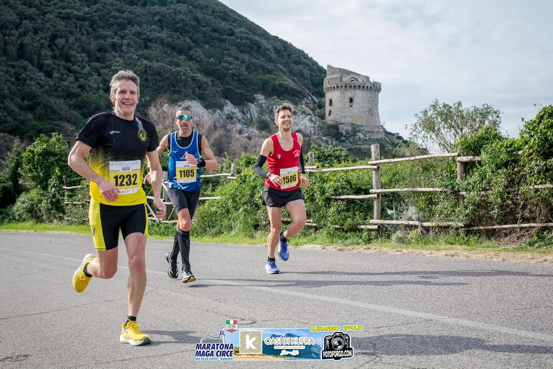 The image shows three people running on a road, likely participating in a marathon or race event. They are wearing bib numbers, and there is a picturesque landscape in the background with hills and a historic-looking tower. The foreground has logos for "Maratona Maga Circe" and other event-related graphics.