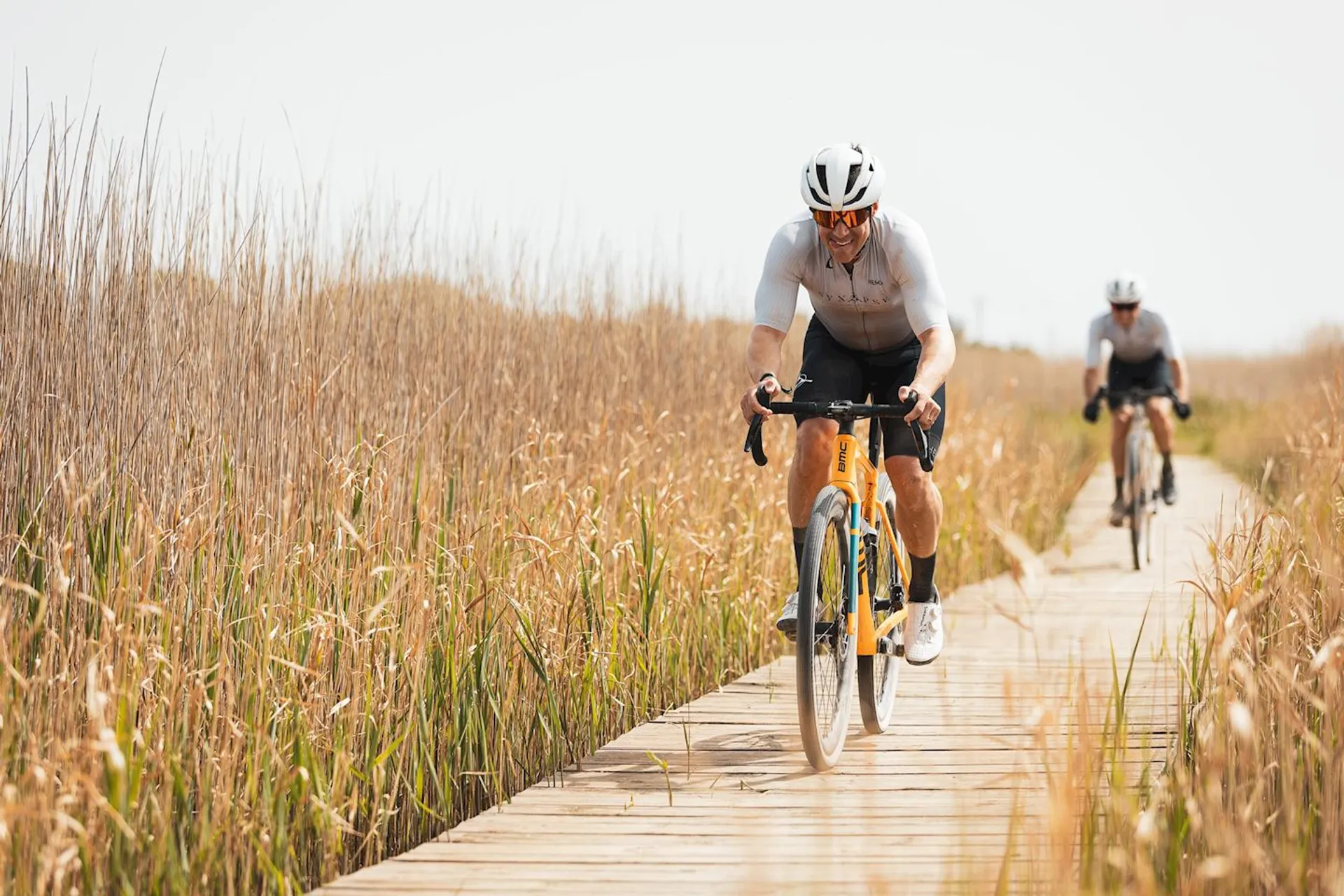 The image shows two cyclists riding on a wooden boardwalk through an area with tall grasses or reeds on either side. The cyclist in the foreground is wearing a white helmet, sunglasses, and a light-colored top, and is riding a road bike with attention focused on the path ahead. The second cyclist is further back and is also seemingly enjoying the ride on a similar pathway. The environment suggests a natural setting, perhaps a marsh or wetland area. The weather appears to be fair, with ample sunlight illuminating the scene.