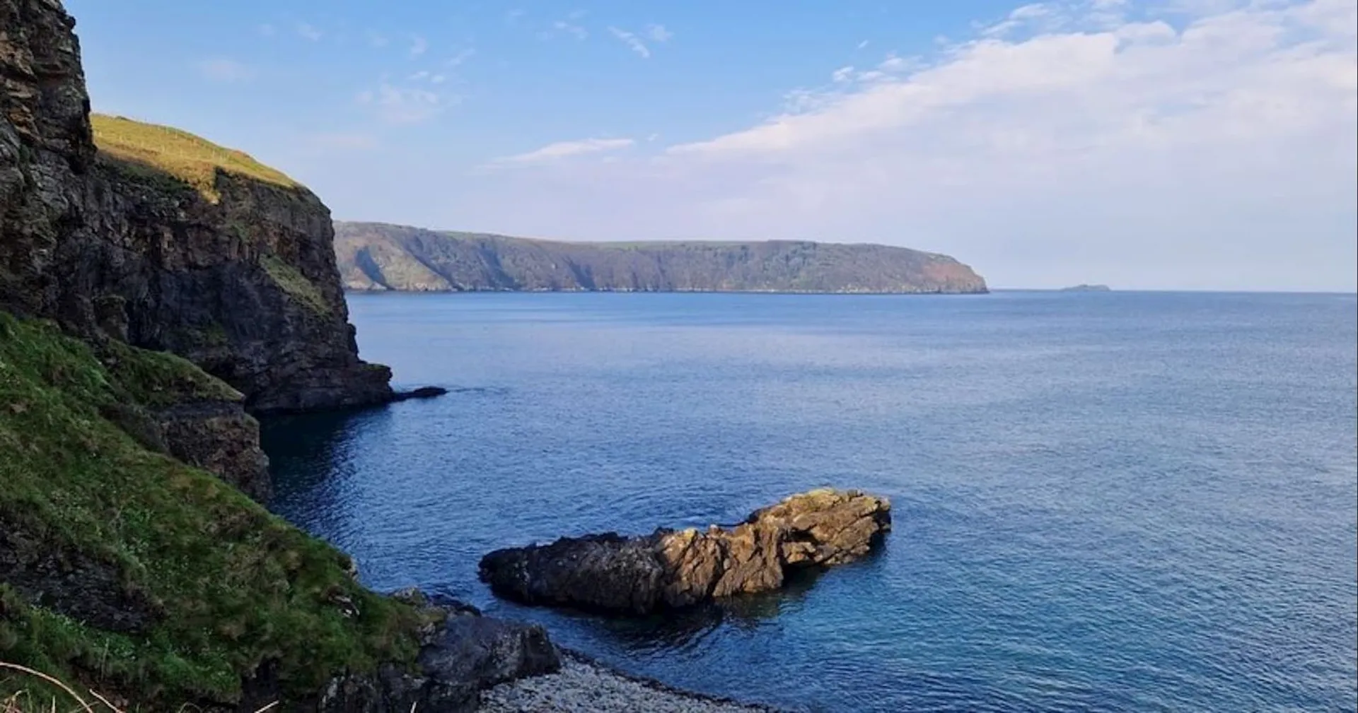 The image shows a coastal scenery. There is a large body of water that appears to be an ocean or sea, with a clear blue sky above it featuring few clouds. On the left side of the photo, there is a steep cliff face leading down to the water. In the distance, you can see additional cliffs stretching along the coastline, suggesting this might be part of a larger coastal region with similar geological features. There's a headland in the distance. The water is calm, and the colors suggest it might be a sunny day. This image captures a serene and natural landscape, likely a place that is visited for its scenic beauty and tranquility.