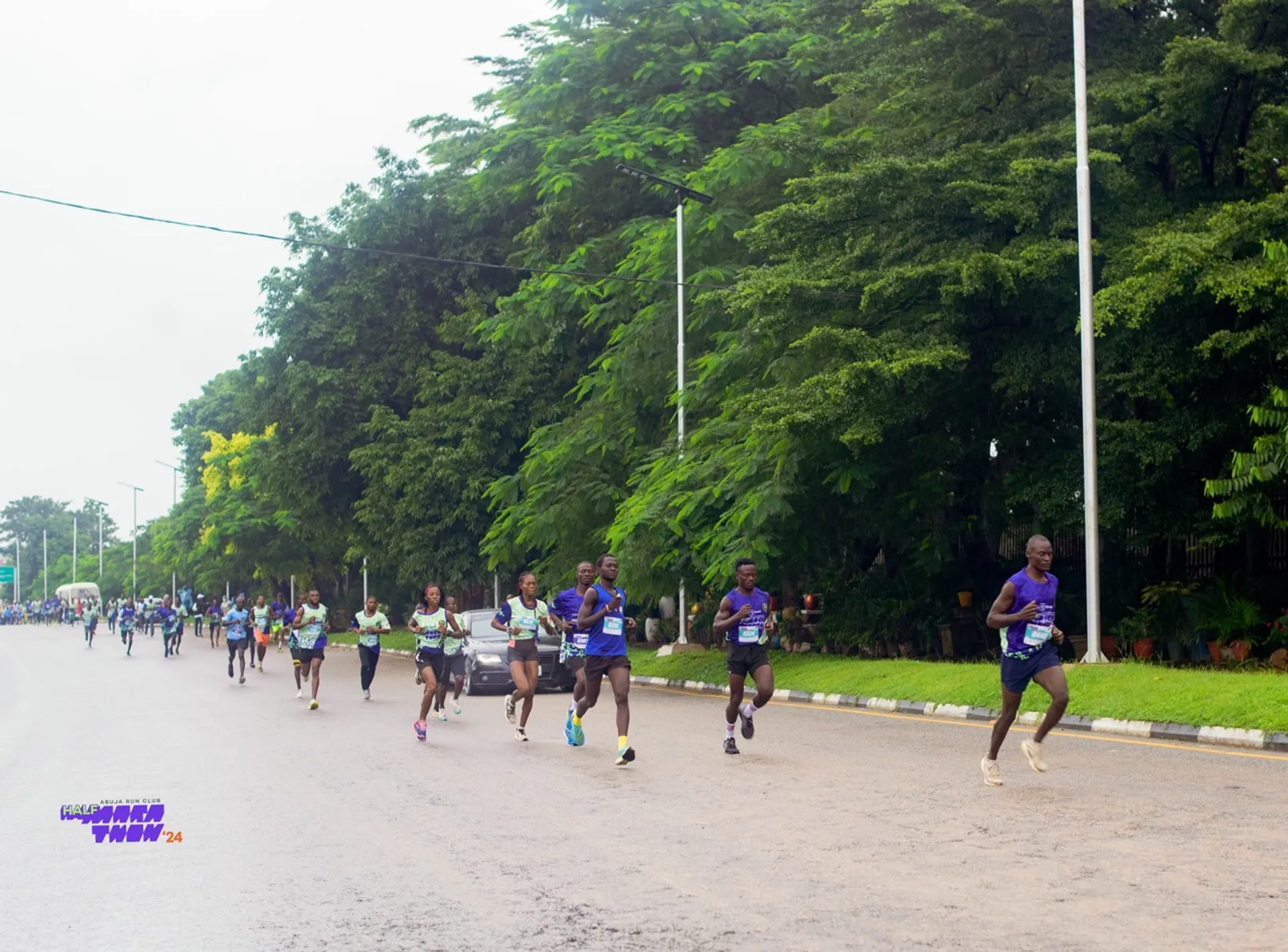The image shows a group of people running in a marathon on a wide street. There are several runners visible, and they appear to be participating in an organized race. The surrounding area is lined with trees and there are streetlights along the road. The sky is overcast, suggesting it might be a cloudy day.