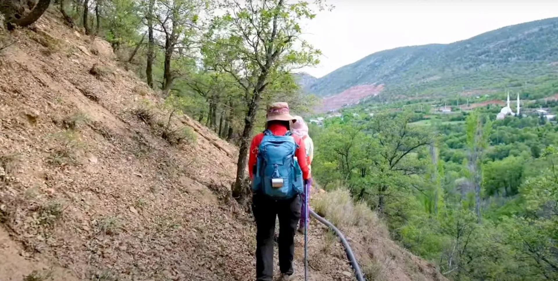 In the image, you see a person from behind, hiking on a narrow dirt trail on a hillside. The hiker is wearing a hat, a backpack, and it seems that they may have a child in a carrier attached to the backpack. In the distance, there is a lush valley with green trees, and further back, you can see a small white building with two minaret-like structures, which could be a mosque. The environment suggests a rural or mountainous area, and overall, it depicts a moment of outdoor adventure or travel.