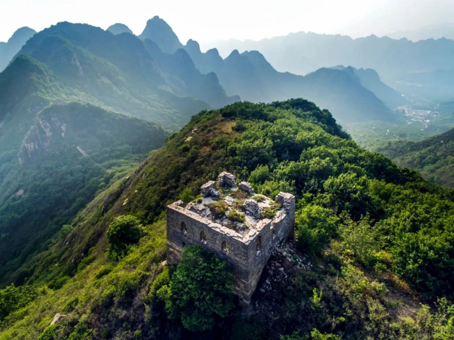 The image shows a scenic view of a dilapidated watchtower on the Great Wall of China, set against a backdrop of lush greenery and mountainous terrain. The structure appears to be overgrown with vegetation, and the mountains in the background are shrouded in mist, creating a dramatic and atmospheric scene.