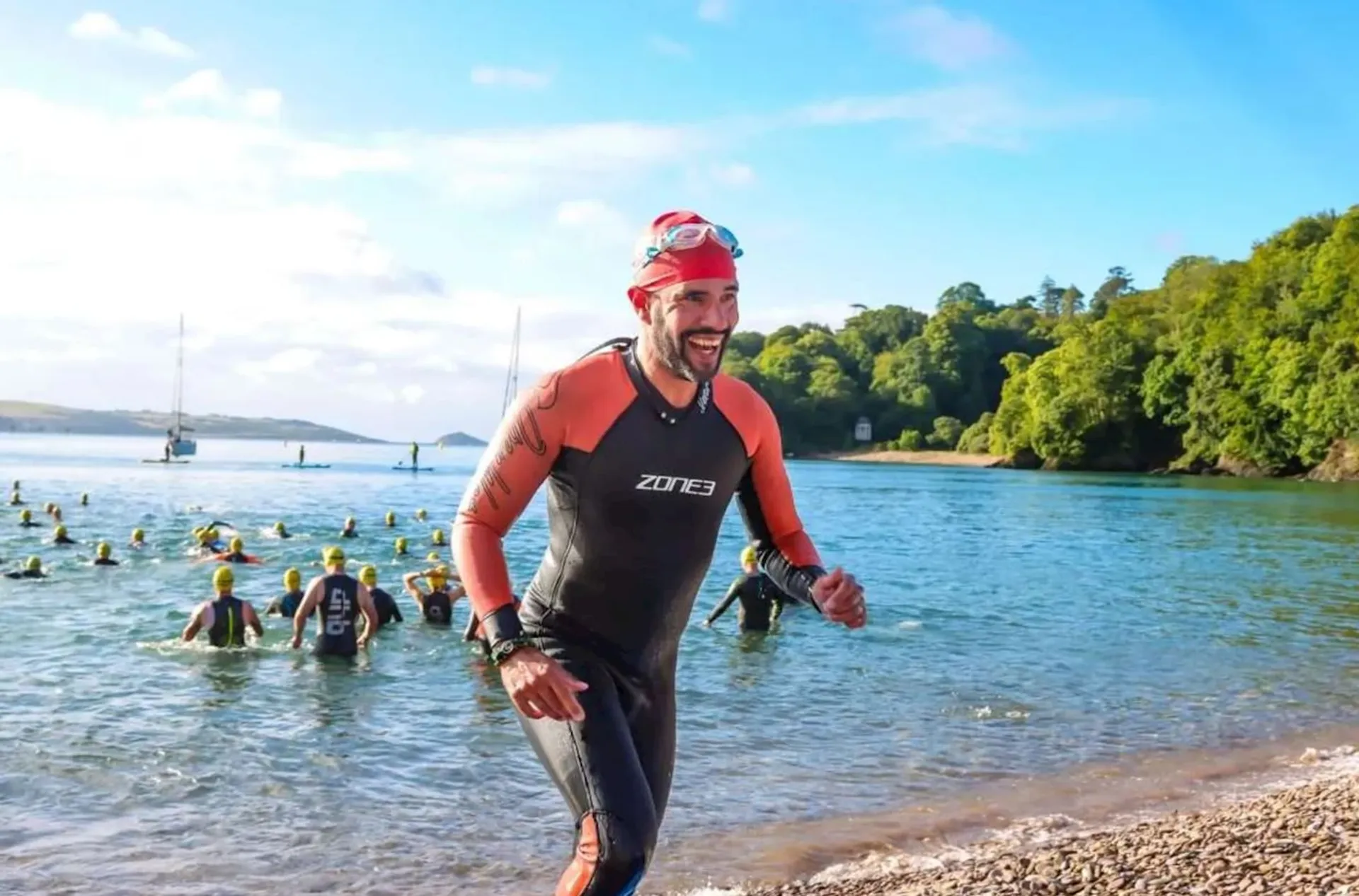 The image shows a person coming out of the water during what appears to be a triathlon or open water swimming event. The individual is wearing a swim cap, goggles, and a wetsuit with the brand "Zone3" visible on it, indicating that they were likely participating in the swimming portion of the event. In the background, you can see other participants still in the water, with some heading towards the shore. The environment suggests that this event is taking place in a natural body of water, possibly a lake or a calm bay, as there are trees on the opposite shore under a clear blue sky, and several sailboats are anchored in the distance. The participants and the setting suggest a friendly and competitive atmosphere on what looks to be a