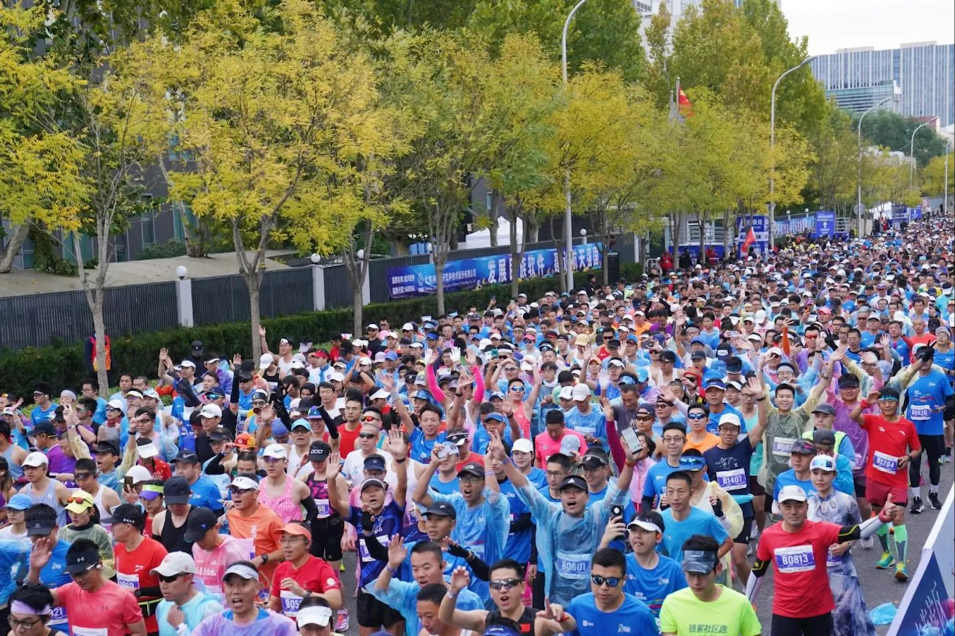 The image shows a large crowd of runners participating in a marathon or large running event on a city street. They're wearing athletic gear, hats, and bibs. Trees line the street, and some banners and spectators are visible in the background.