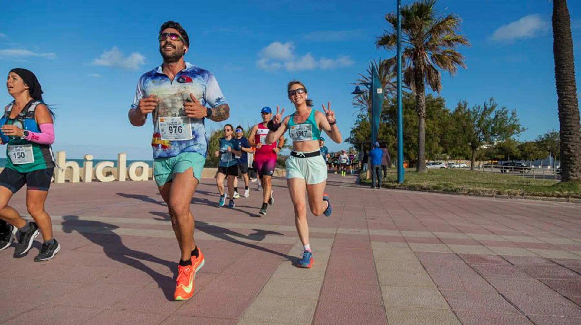 The image shows a group of people running in what appears to be a race or marathon. They're on a paved path with palm trees and a clear sky in the background. Some runners have bib numbers, and the environment looks bright and sunny.