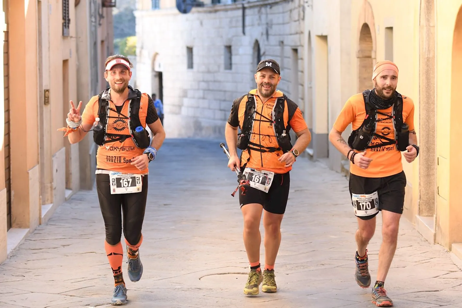 The image shows three individuals running down a stone-paved street, likely participating in