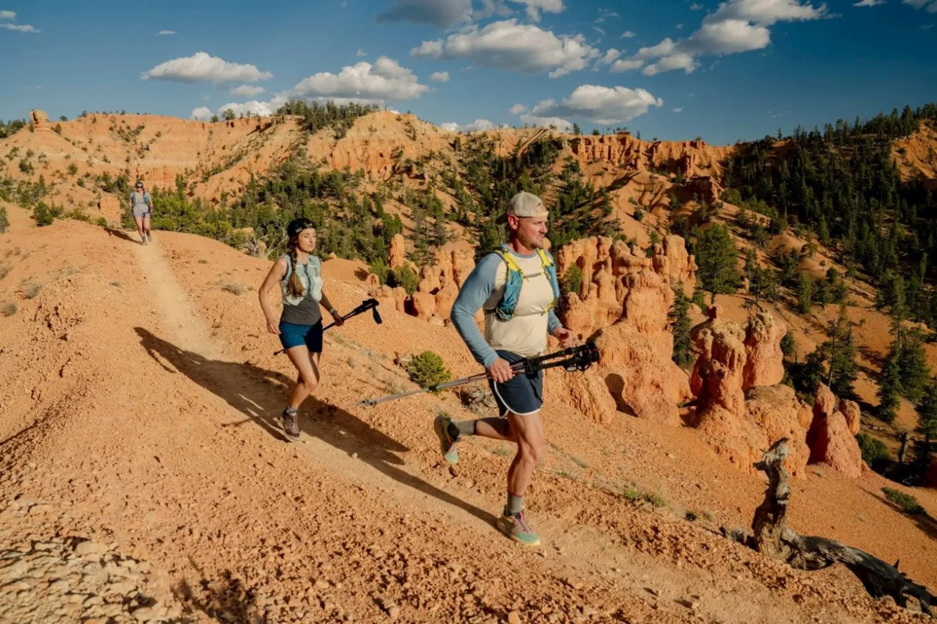 The image shows a group of people trail running in a desert landscape. They are on a dirt path surrounded by rocky formations and sparse vegetation typical of a desert environment. The sky is blue with some clouds. The scene appears to be outdoors in a natural, rugged setting.